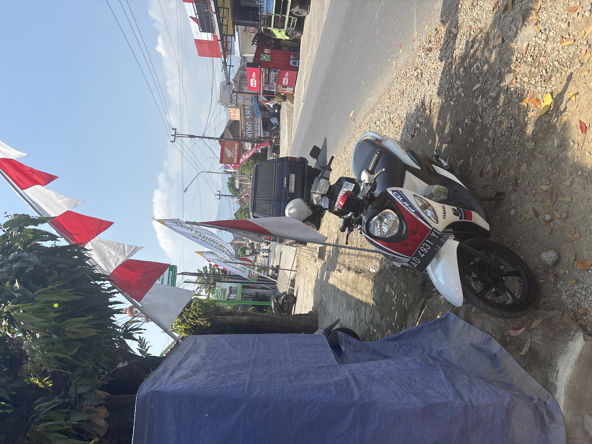 This image depicts a street scene in Tirtomoyo, Indonesia, characterized by the presence of Indonesian flags and a motorcycle parked on the side of the road. A blue tarp partially obstructs the left side of the frame, suggesting a temporary structure or covering. Numerous red and white Indonesian flags are strung along the street, fluttering in the breeze, with one particularly prominent flag in the foreground.
A white and red scooter, a Yamaha Fino, is parked on the gravel shoulder, its license plate reading "AD 4931 YI". A white helmet rests on the handlebars. Behind the scooter, a dark blue SUV with license plate "AD 1292 KY" is parked parallel to the road.
The street itself is paved, with a sidewalk on the right side where various shops and buildings are visible. Signs in Indonesian are present, including one for "Honda" and another for "BeNice Coffee". A green truck is parked in front of one of the buildings. The environment is sunny, with clear blue skies and some scattered clouds. The ground is covered in gravel and dry leaves, indicating a dry season. A few people are visible in the background, walking or riding motorcycles. The overall impression is of a typical Indonesian street scene, possibly during a national celebration given the abundance of flags.