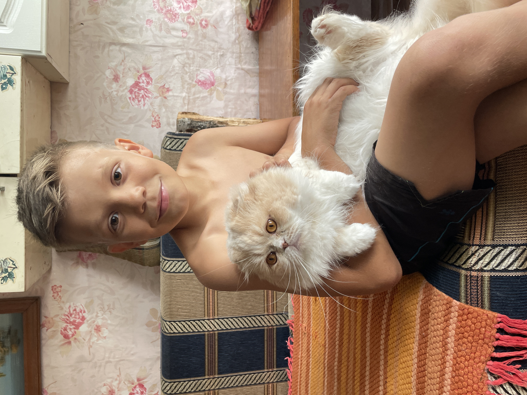 A young boy is sitting on a striped couch, holding a fluffy cat. The boy is shirtless and wearing black shorts. The cat is cream and white with orange eyes. The couch has blue, beige, and brown stripes. To the left of the couch is a woven blanket with orange and yellow stripes and red fringe. Behind the boy is a wall with floral wallpaper and a wooden cabinet. A wooden table is visible to the right. The setting appears to be indoors, in a home. The image was taken in Маминское, Russia.