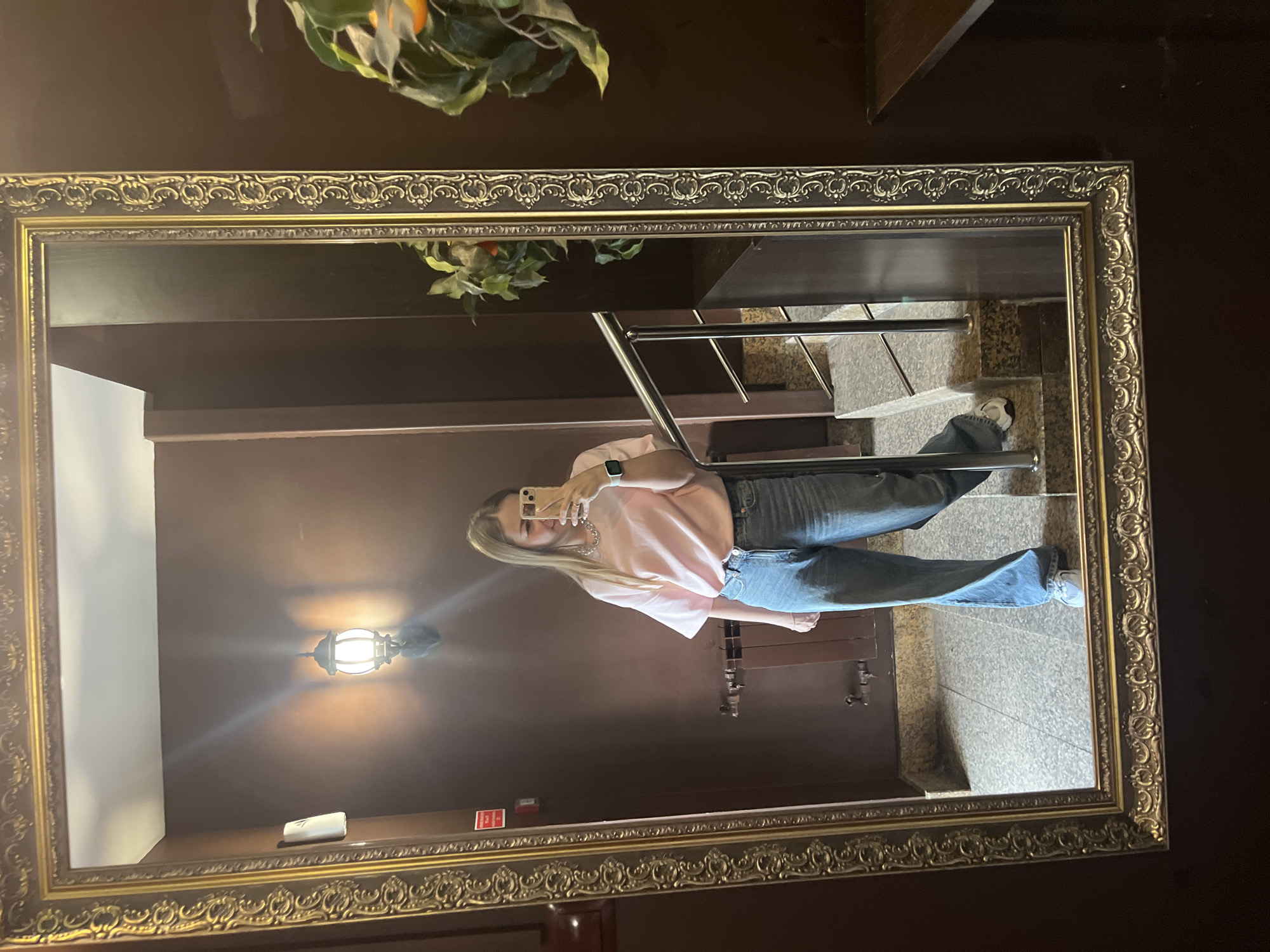 A young woman is depicted taking a mirror selfie in an indoor setting. She has long, straight, light-colored hair and is wearing a pink oversized short-sleeved t-shirt, light blue wide-leg jeans, and white sneakers. On her left wrist, she wears a smartwatch with a green band, and a silver chain necklace is visible around her neck. She holds a light-colored smartphone with both hands, positioned to capture her reflection.
The mirror she is using has a thick, ornate, gold-colored frame and is mounted vertically on a dark brown wall. Reflected in the mirror, behind the woman, is a dimly lit hallway or stairwell. The walls are consistently dark brown. A wall-mounted lantern with a warm, glowing light source is visible on the upper left side of the reflection, casting light onto the dark wall. Above the lantern, a white rectangular device is affixed to the wall, and a small red sign with white lettering (partially obscured) is present lower on the left. Below these, a dark-colored radiator or heating unit is visible.
To the woman's right, a set of stairs with light grey, speckled granite-like treads and risers ascend upwards. A modern metal railing with silver posts and horizontal bars runs alongside the stairs. The floor area visible at the base of the stairs and near the woman's feet is also covered in the same light grey, speckled tile. A faux plant with green leaves and orange spherical fruit (resembling tangerines or small oranges) is reflected on the far right side of the mirror, suggesting it's situated outside the immediate mirror frame but within the room. Another small, dark plant is visible lower on the left side of the mirror's reflection. The overall environment suggests a private or semi-public interior space, possibly a lobby, corridor, or stairwell in a building. The city and specific location of this scene are unknown.