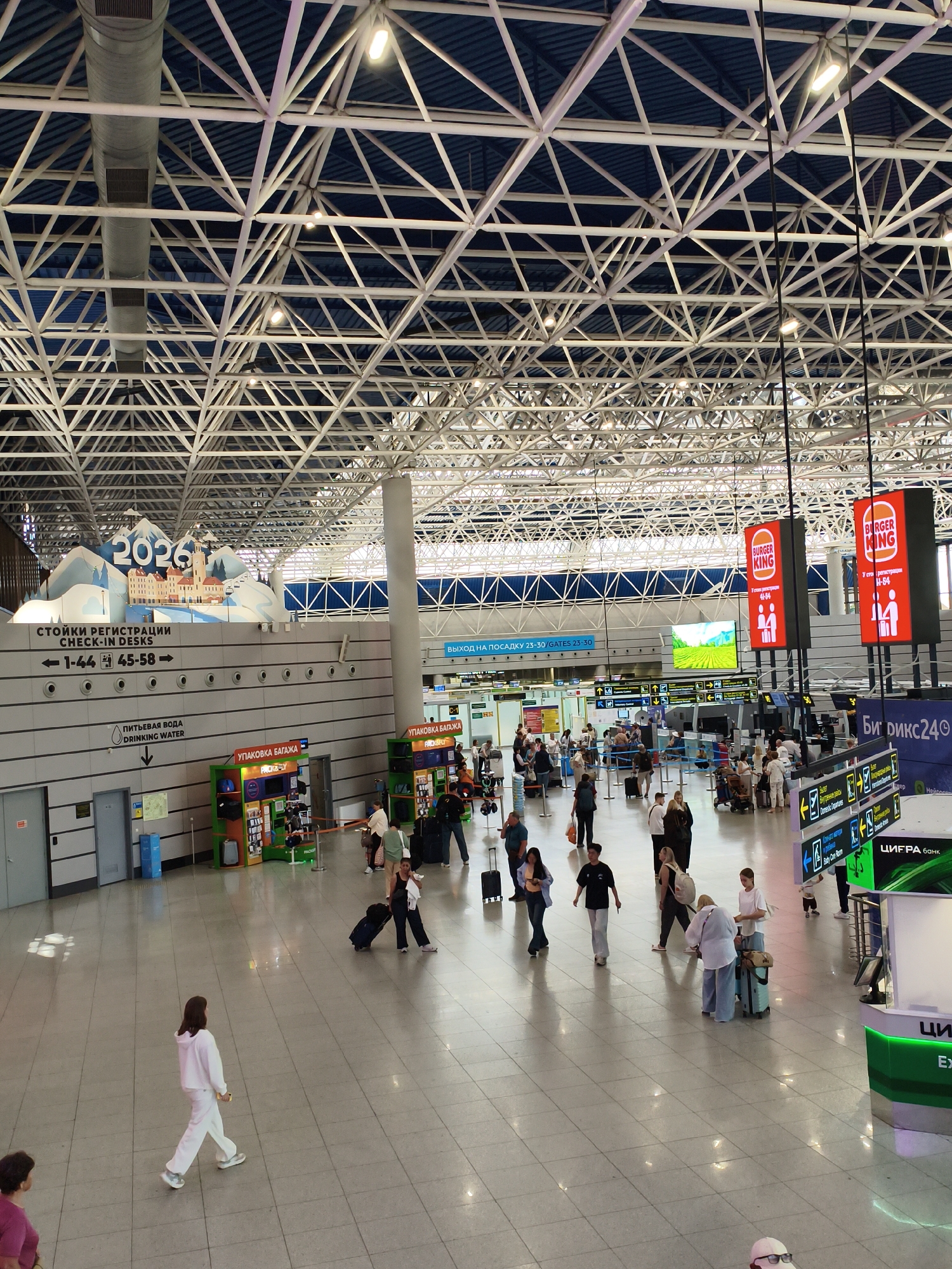 This is a photograph of the interior of the Sochi International Airport in Sochi, Russia. The image captures a wide view of the departure hall, characterized by its high, white, lattice-like ceiling structure and vast, tiled floor.
In the foreground, several individuals are walking through the space, some with luggage, indicating travel activities. A woman in a white tracksuit walks away from the camera, while other figures are seen in the mid-ground, some interacting with check-in counters or waiting in lines.
Prominently displayed in the background are large digital signs for Burger King and informational boards indicating check-in desks and gates. A large decorative banner with "2026" and mountain imagery is visible above the check-in area, likely related to the Olympic Games. The environment is well-lit, with artificial lighting and natural light filtering through unseen sources. The overall scene depicts a busy airport terminal in operation.
