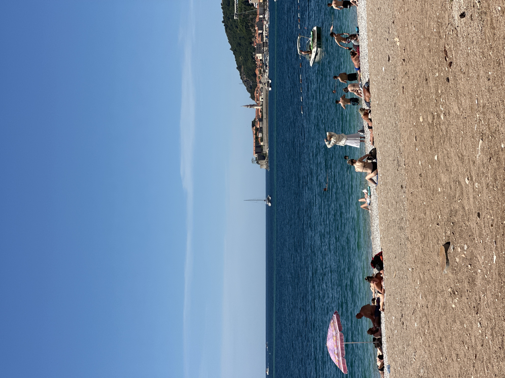 The image shows a beach in Budva, Montenegro, on a sunny day. People are swimming and relaxing on the beach, a coastal town nestled along the Adriatic Sea. A boat is visible in the water. A line of buildings, including a church steeple, lines the coast. The sky is blue, and the water is a deep blue color. A beach umbrella is partially visible at the bottom of the image. The beach consists of pebbles and sand.