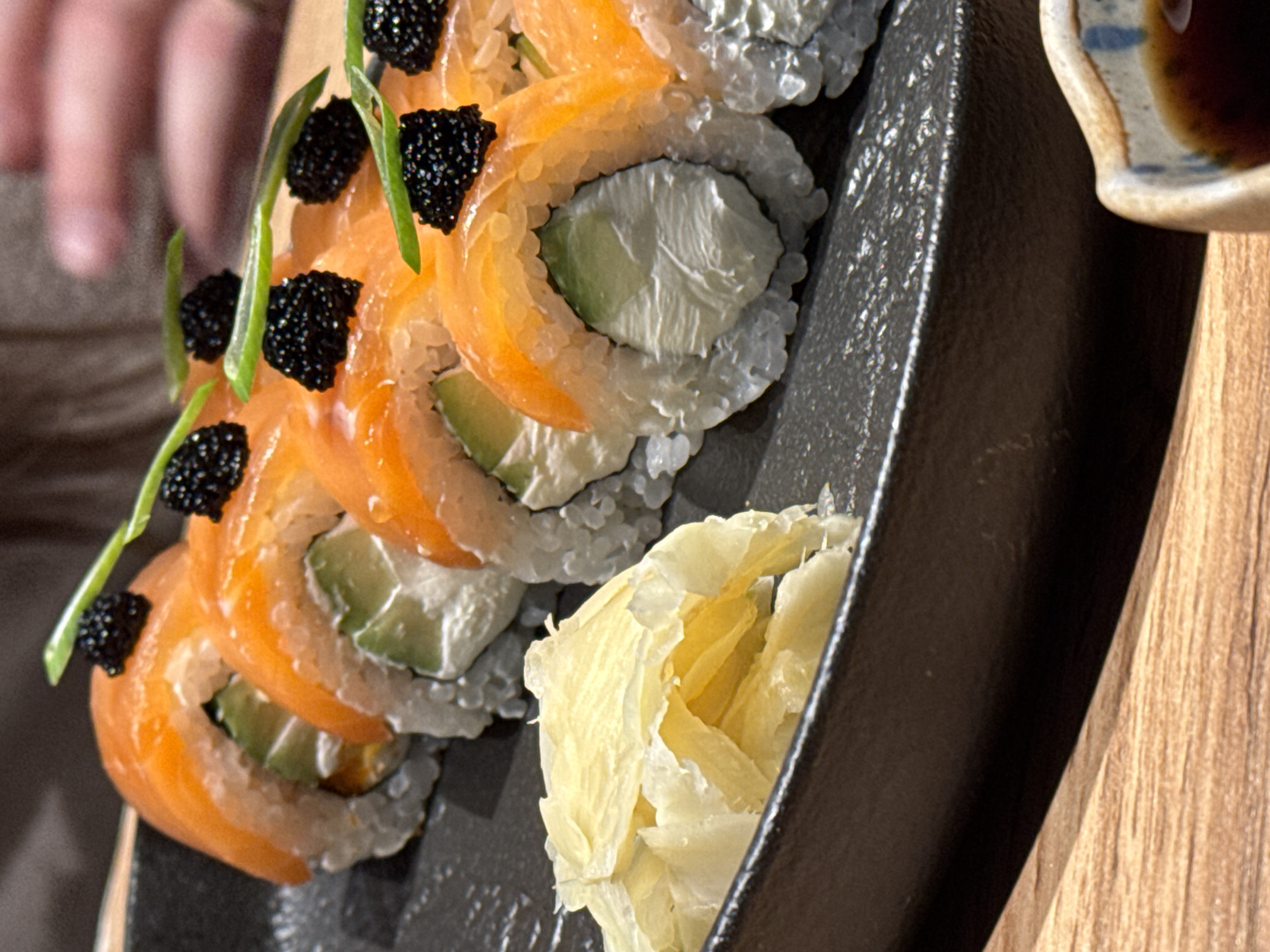 A close-up shot shows a row of sushi rolls on a black plate, topped with salmon, black caviar, and thin green sprigs. The rolls appear to contain rice, avocado, and possibly cream cheese. Pickled ginger is also on the plate. A small dish containing a dark liquid, likely soy sauce, is partially visible at the right edge. The plate rests on a wooden surface. The scene appears indoors, possibly in a restaurant. The photograph was taken at a restaurant named "Khmeli-Suneli" in Chaikovsky, Russia.