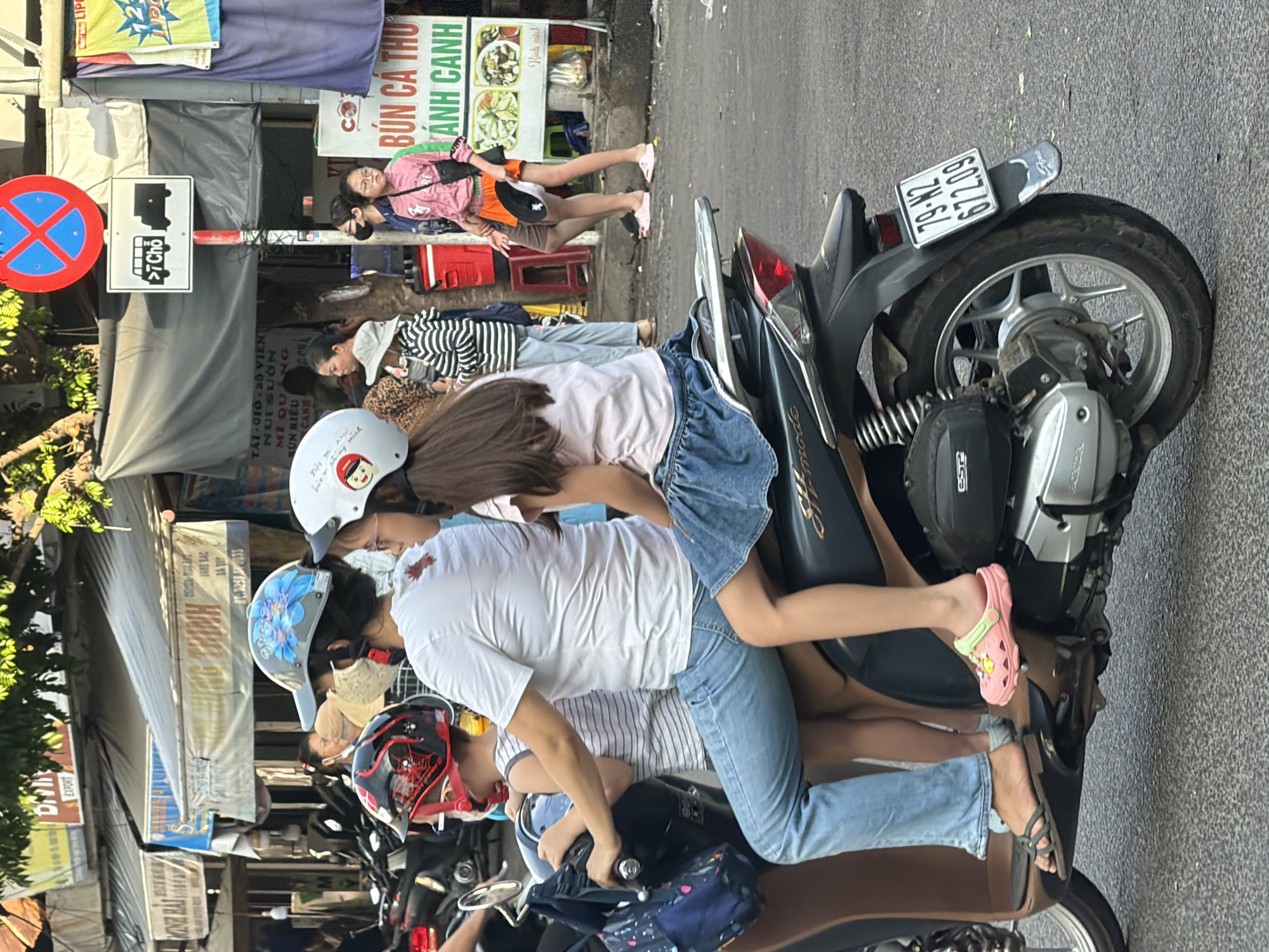 The image depicts a street scene in Nha Trang, Vietnam, likely on a bright day. In the foreground, a family of three is riding a scooter. A woman, wearing a white t-shirt and blue jeans, is driving the scooter. A young girl, with long brown hair and wearing a white helmet, sits behind her, dressed in a pink top and blue denim skirt. In front of the woman, a child is seated, wearing a black and red helmet and a striped tank top. The scooter has a license plate with the characters "79-N2 672.09". The background shows a bustling street with various shops and awnings. Signs with Vietnamese text are visible, including one that reads "BÚN CÁ THU ANH CANH". Several other people are walking and standing in the background, and a number of other motorcycles are parked. The overall atmosphere suggests a typical day in a Vietnamese city.