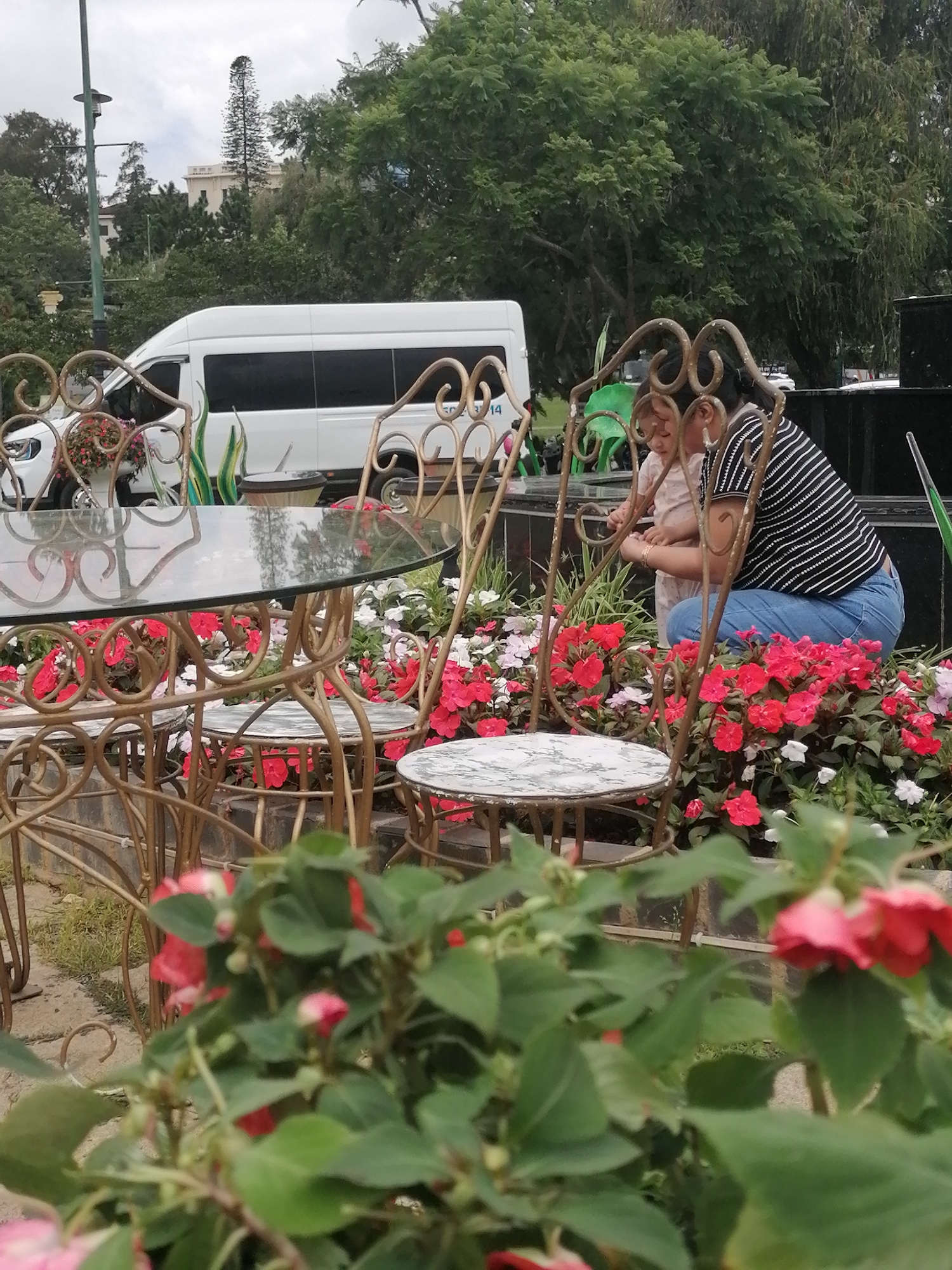 In Da Lat, Vietnam, a woman in a black and white striped shirt and blue jeans crouches down next to a young child. They are seated in front of a floral arrangement of red and white impatiens, surrounded by ornate golden metal garden furniture, including a table with a glass top and two chairs. In the background, a white minivan is parked near a lush green treescape and a building.