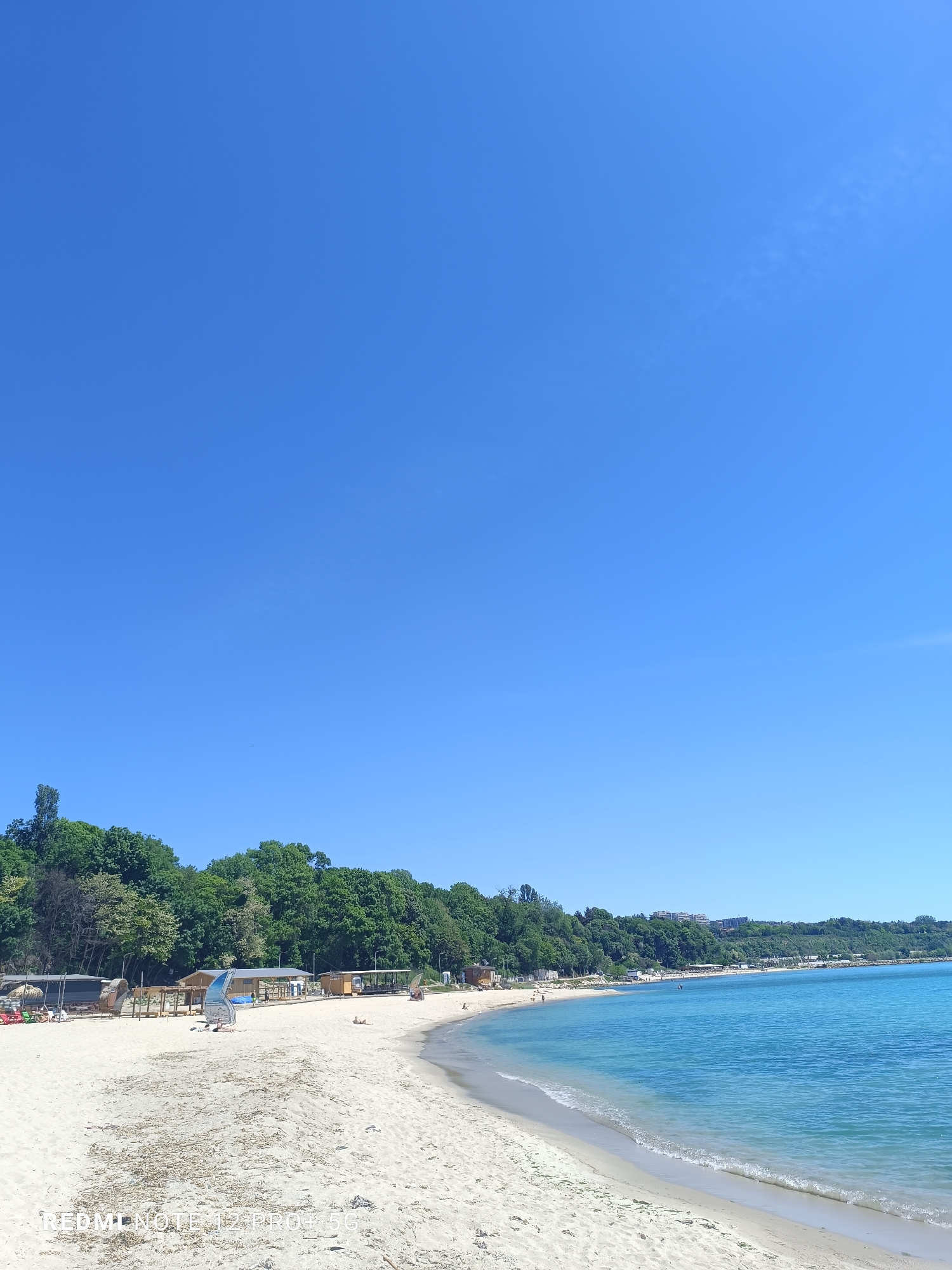 The image depicts a beach on a sunny day. The sky is clear and blue. A sandy beach stretches from left to right, with a line of trees bordering it at the back. The water is blue and clear. There are a few buildings visible in the background behind the trees. The location is likely a beach near the city of Varna, Bulgaria. There are a few people on the beach, some of whom are sitting or lying down. A windsurf is on the beach.