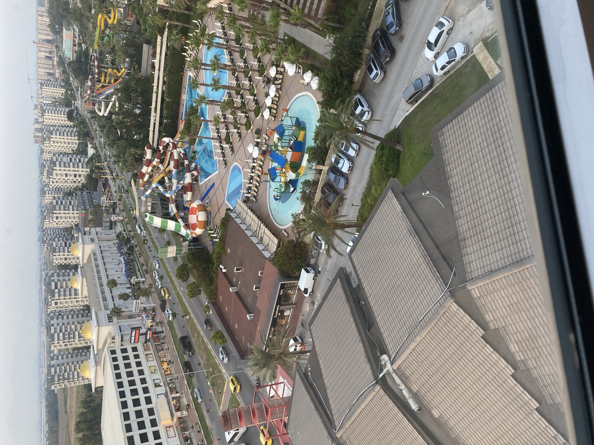 An aerial perspective captures a scene in Aksu, Turkey, likely a resort area given the presence of a waterpark. Multiple multi-story apartment buildings dominate the skyline, interspersed with trees and vegetation. A large shopping mall, "Hisar Mall," is visible, with vehicles on the road in front of it. The waterpark features several slides, pools, and lounge chairs. Rows of parked cars are scattered throughout the area, and several white vans are present. The foreground is dominated by the tiled roofs of buildings. The sky is overcast.