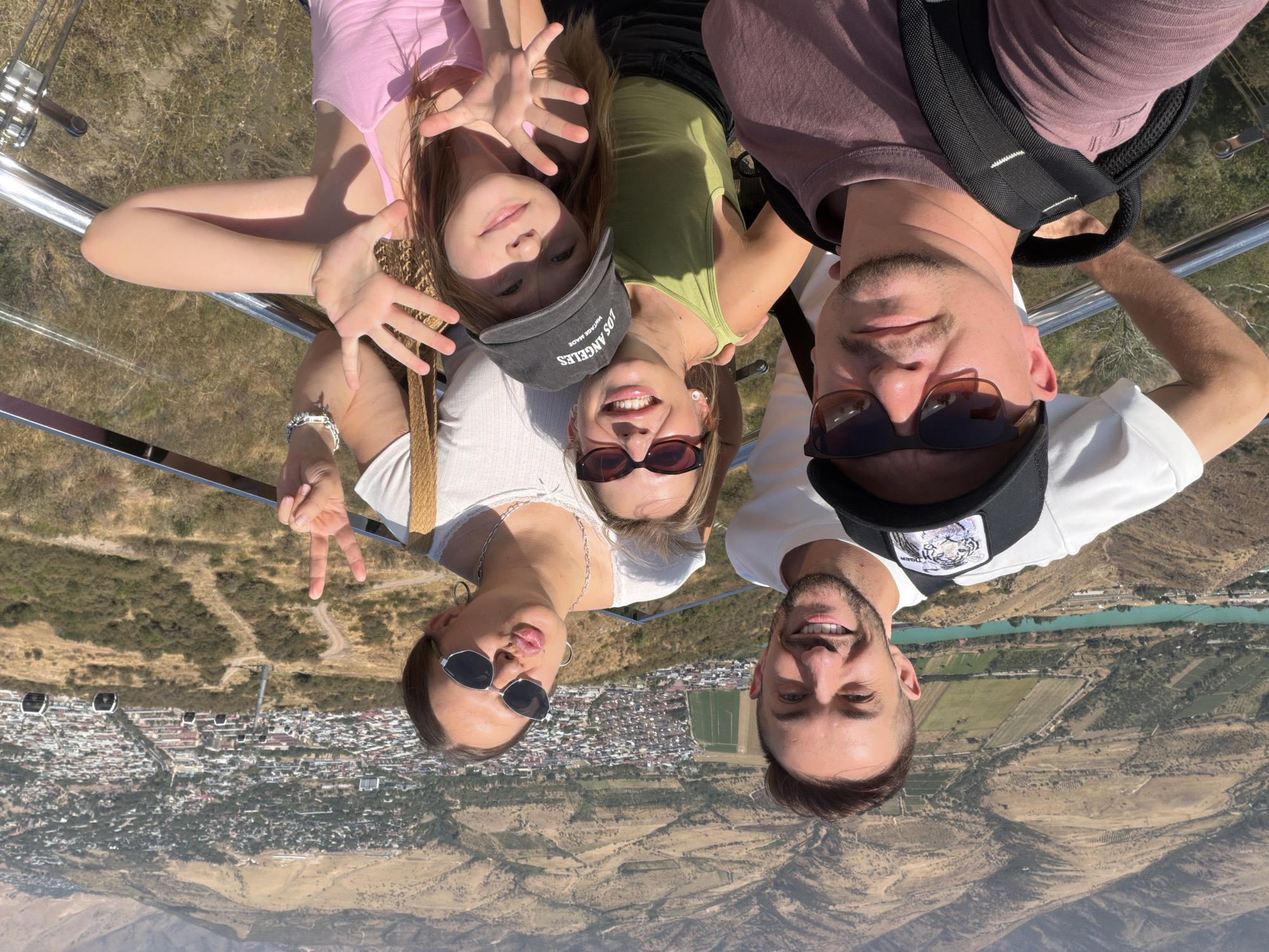 This is a selfie of five people standing on a viewpoint with a city and mountains in the background. The people are arranged in two rows, with three in the back and two in the front. The person on the far left in the back is a man with short dark hair, a beard, and a white t-shirt, smiling at the camera. To his right, in the back row, is a woman with dark hair, sunglasses, and a white top, puckering her lips and holding up two fingers in a peace sign. Next to her, also in the back row, is another woman with blonde hair, sunglasses, and a green tank top, smiling. In the front row, on the left, is a man wearing sunglasses, a baseball cap with a tiger design, and a purple t-shirt, looking at the camera. To his right is a young girl with long red hair wearing a pink tank top and a grey baseball cap that says "Los Angeles," smiling and holding up her hands. They appear to be at a scenic overlook in the city of Charvak, Uzbekistan, with a valley and river visible below, as well as cable cars in the distance. The weather is sunny and clear.
