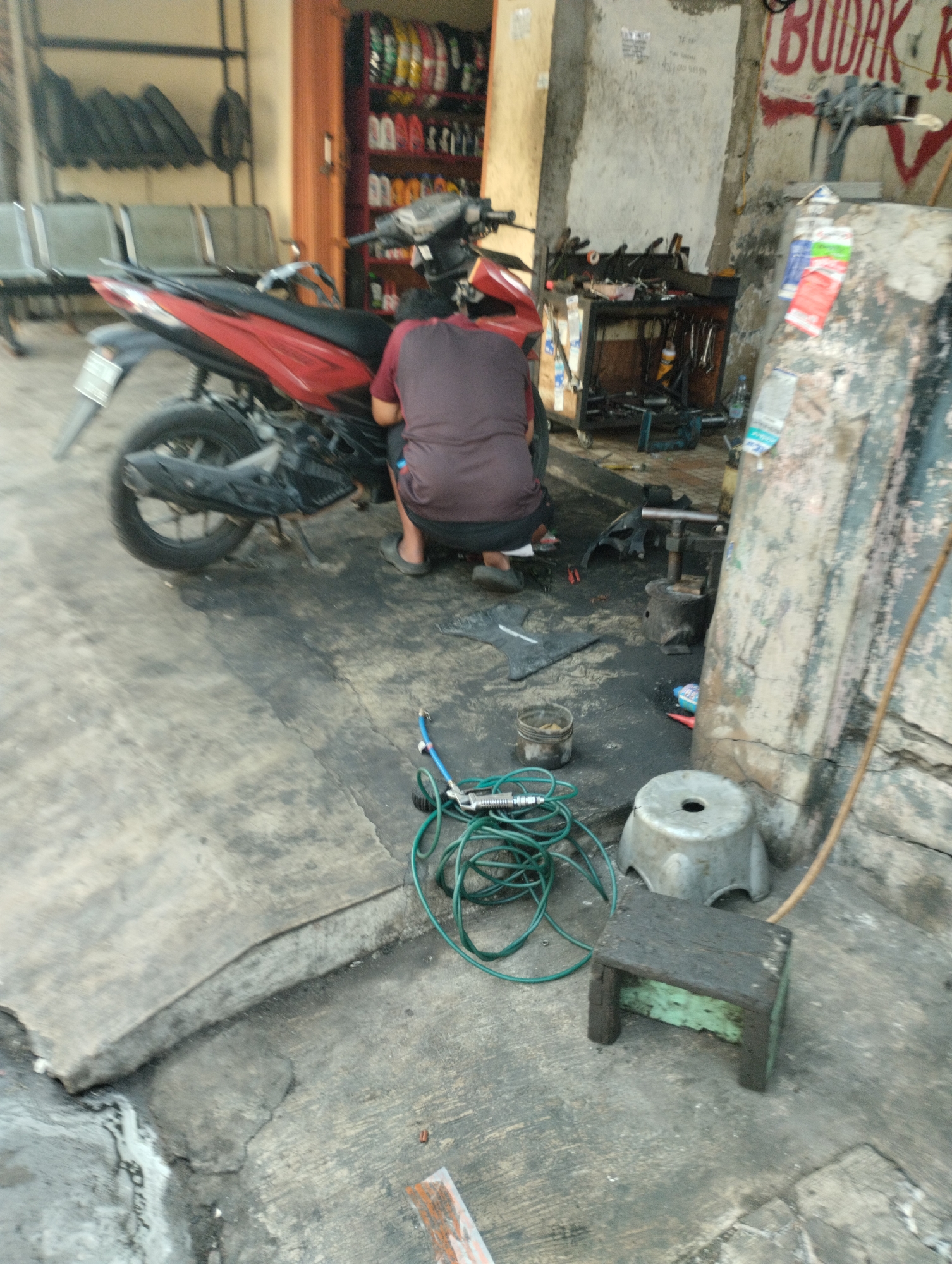 The image depicts a person working on a red and black motorcycle in what appears to be a roadside workshop in Bekasi, Indonesia. The workshop is cluttered, with various tools and parts visible on the ground and on shelves. A display of tires and other motorcycle accessories is seen in the background. The ground is concrete, with some oil stains and debris scattered around. The lighting suggests it is daytime, possibly late afternoon, with a somewhat hazy or overcast sky. The person, wearing a maroon t-shirt and dark shorts, is crouched beside the motorcycle, seemingly focused on a repair. A green hose and a metal tool are lying on the ground near the person. The overall scene conveys a sense of everyday work in a typical urban setting in Indonesia.
