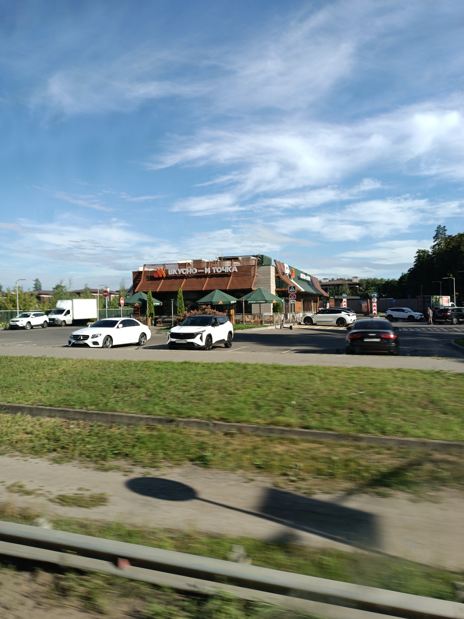The image shows a "Vkusno & tochka" (formerly McDonald's) restaurant in Malakhovka, Russia, on a bright, sunny day with a blue sky and scattered clouds. Several cars are parked in the lot and driving through. In the foreground, a grassy area with a concrete border runs along a road, partially obscuring the view with motion blur. The "Vkusno & tochka" building has a distinctive brown and green color scheme with awnings over the windows. A white delivery truck is parked to the left of the building. The scene suggests a typical day at a fast-food restaurant.