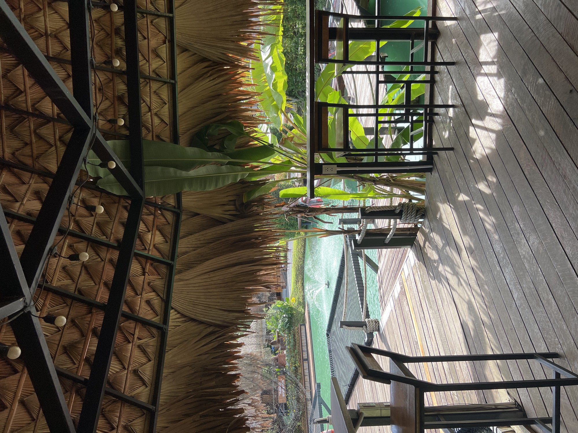 The image depicts an outdoor seating area with a thatched roof, offering a view of a turquoise-colored body of water with a fountain. The seating area is constructed with a wooden deck and features high tables with bar stools. In the background, lush green foliage and some buildings can be seen. The atmosphere suggests a relaxed, possibly resort-like setting. The scene is captured during daylight, with sunlight casting shadows on the wooden deck.