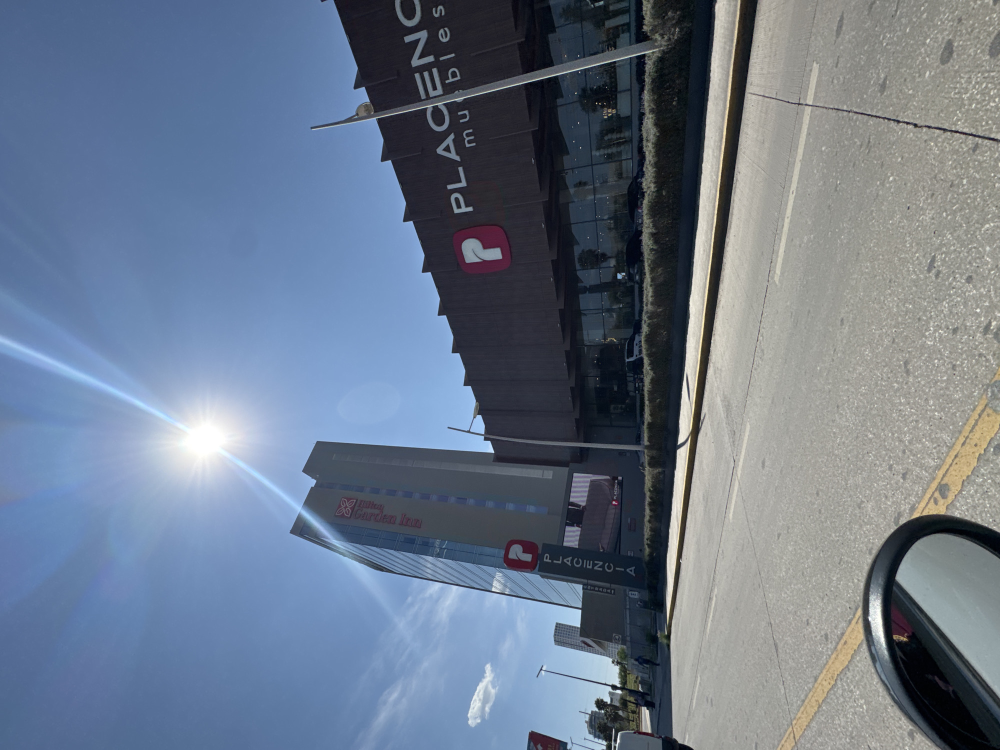 The image shows a street scene in San Bernardino Tlaxcalancingo, Mexico. The sun is high in the sky. On the right side of the image, a portion of the street with yellow markings is visible. On the left are businesses: "Placencia muebles" and a Hilton Garden Inn. A fragment of a side mirror of a vehicle appears on the right. Several people are visible on the street.