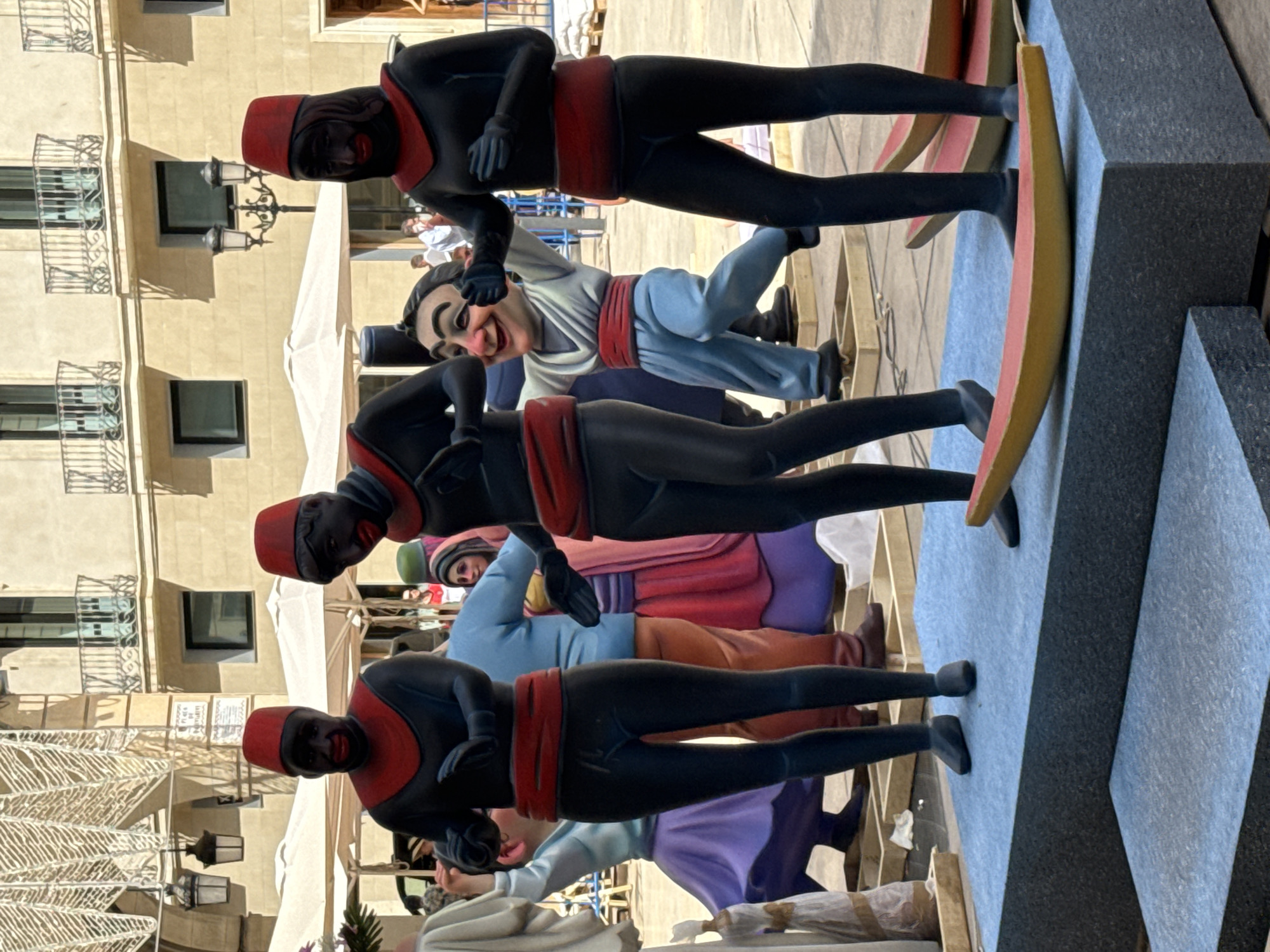 A group of figures, likely part of an outdoor festival display, are positioned on a raised platform in an urban setting. Three of the figures are dressed in black leotards with red sashes and turbans. Their faces are painted black with red lips. Between these figures, a smaller, stylized character with a large, grinning face, wearing a white tunic and blue pants, is also present. In the background, buildings with balconies and awnings are visible, suggesting a public square or street. The ground is paved, and there are white tents and some scaffolding in the distance. The overall scene appears to be set during the daytime, with ample natural light.