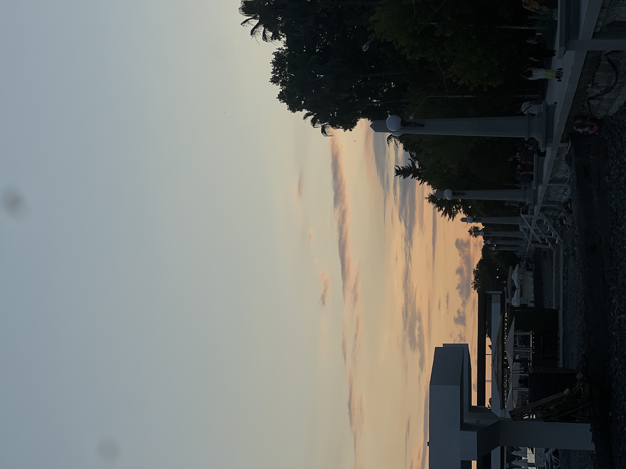 The image depicts a waterfront promenade in Sokhumi, Abkhazia, at dusk. The sky displays a gradient of pale blue to soft orange, with thin, wispy clouds tinted pink and orange by the setting sun. Silhouetted against the colorful sky are palm trees and dense foliage lining the promenade. Several tall, classical-style lampposts are evenly spaced along the paved walkway.
In the foreground to the left, a portion of a white, modern-style structure, possibly a canopy or part of a building, is visible, with some wooden planks leaning against it. Further back, beach umbrellas and cabanas suggest a recreational area. The ground is covered with dark pebbles.
On the promenade, several people are casually walking or standing. One individual is riding a scooter. The overall atmosphere is tranquil, characteristic of an evening stroll along the seaside. The lighting is dim, consistent with twilight.