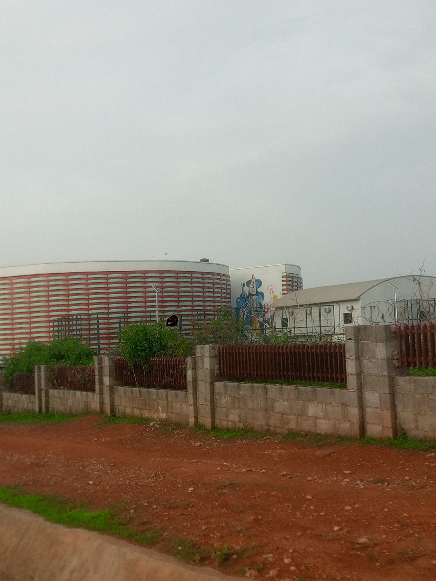This is a medium-distance, eye-level, full shot taken on an overcast day in Gwarinpa, Nigeria. The scene features a large, cylindrical building with a white top and a facade of horizontal red and white stripes. To the right of this building is a smaller, rectangular white building with a curved roof. A mural depicting a person in blue and yellow clothing holding a staff is visible on the side of the larger building. A brown metal fence with vertical slats runs along the foreground, supported by concrete pillars and a low concrete block wall. Behind the fence, lush green bushes and trees are visible. The ground in front of the fence is a mixture of red dirt and scattered small stones, with a strip of green grass along the far left edge of the frame. The sky is a uniform light gray, indicating cloudy weather.