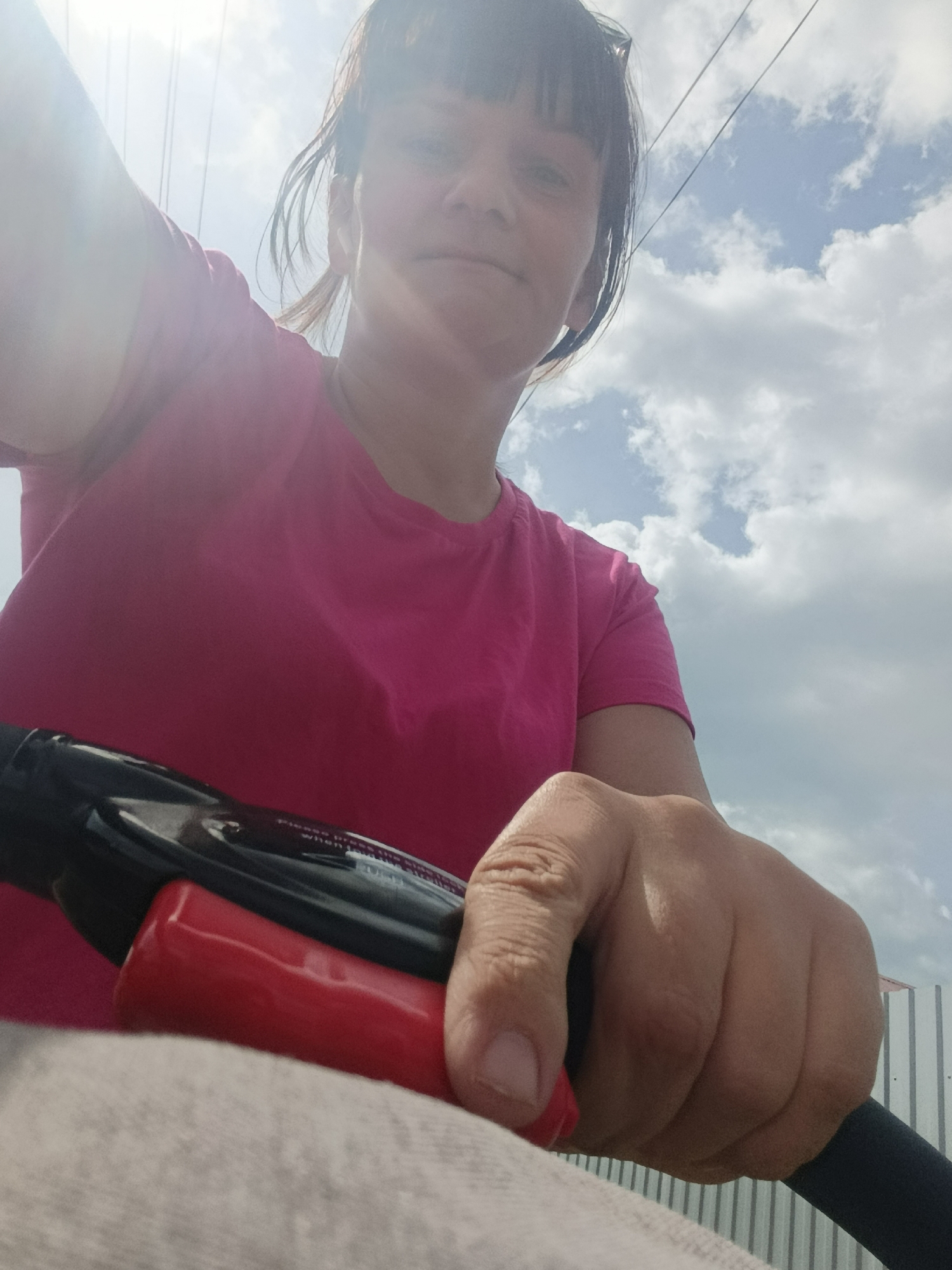 A woman with bangs and a pink shirt is holding a black and red handle, likely part of a bicycle, on what appears to be a partly cloudy day. The photo is taken from a low angle, looking up at the woman and the sky. Power lines are visible in the upper portion of the image. In the background, a corrugated metal fence can be seen. The specific location in Фоки, Russia, cannot be determined from the image.