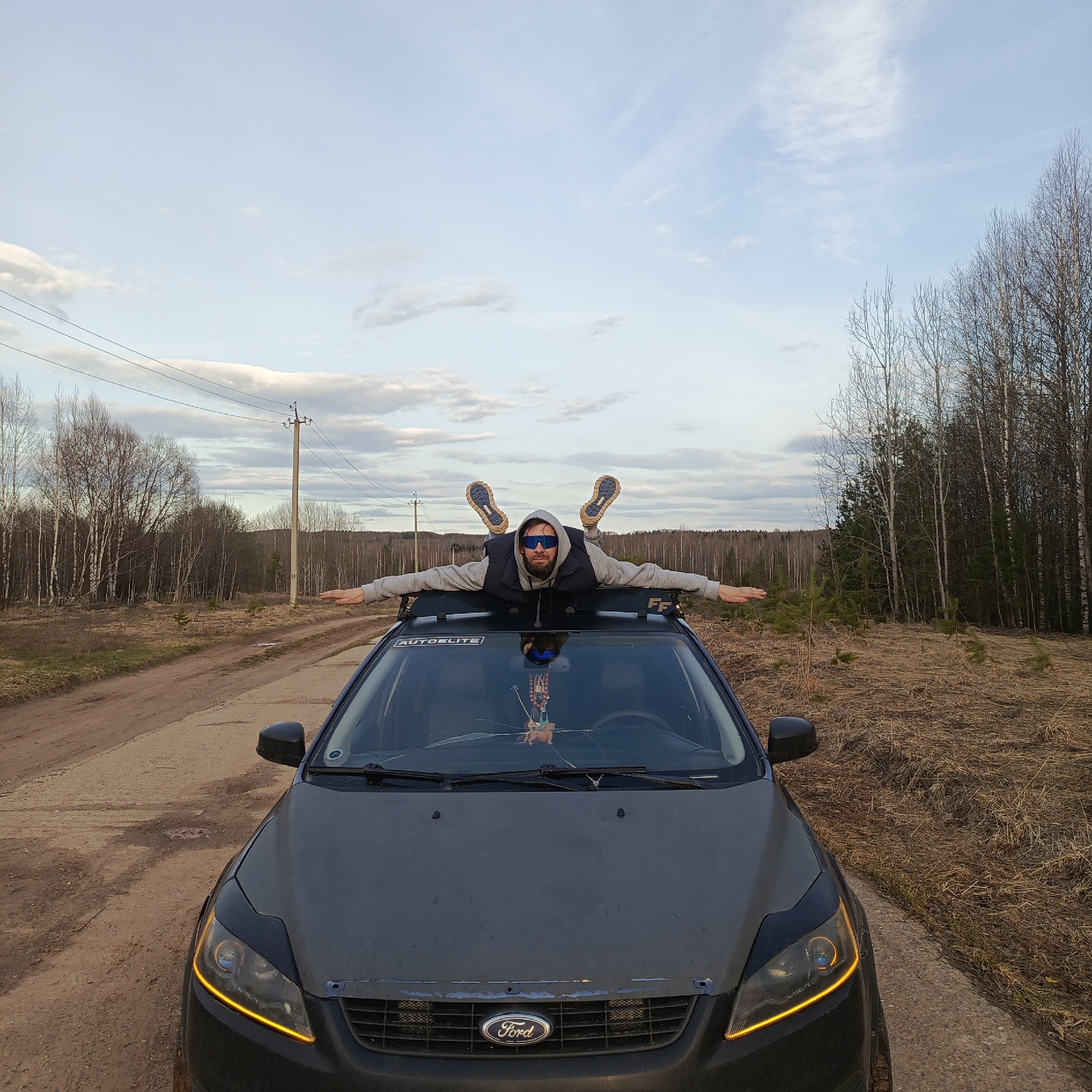 A man is lying flat on the roof of a dark grey Ford Focus on a dirt road in a rural area. The man is wearing a grey hooded sweatshirt, a black vest, sunglasses, and hiking boots. His arms are outstretched, and his legs are raised in the air. The car has a roof rack and aftermarket headlights. The windshield is cracked. The background consists of a dirt road, dry vegetation, bare trees, a few evergreen trees, and utility poles. The sky is partly cloudy. The location is Марковский, Russia.