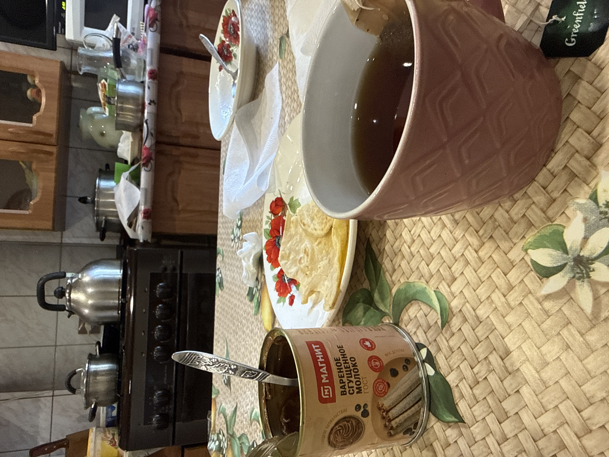 A close-up, high-angle shot captures a kitchen table setting in Valday, Russia, featuring a meal and beverages. The table is covered with a woven mat adorned with a floral pattern. In the foreground, a pink mug with a textured design holds dark tea with a tea bag tag that reads "Greenfield." To the left of the mug, an open can of condensed milk, labeled in Russian as "Magnit Varenoe Sgushchennoe Moloko," is visible with a spoon inside. Adjacent to the can is a plate with thin, golden crisps. In the background, a stove with pots and kettles is partially visible, suggesting a domestic kitchen environment. The overall scene depicts a casual moment of consumption.