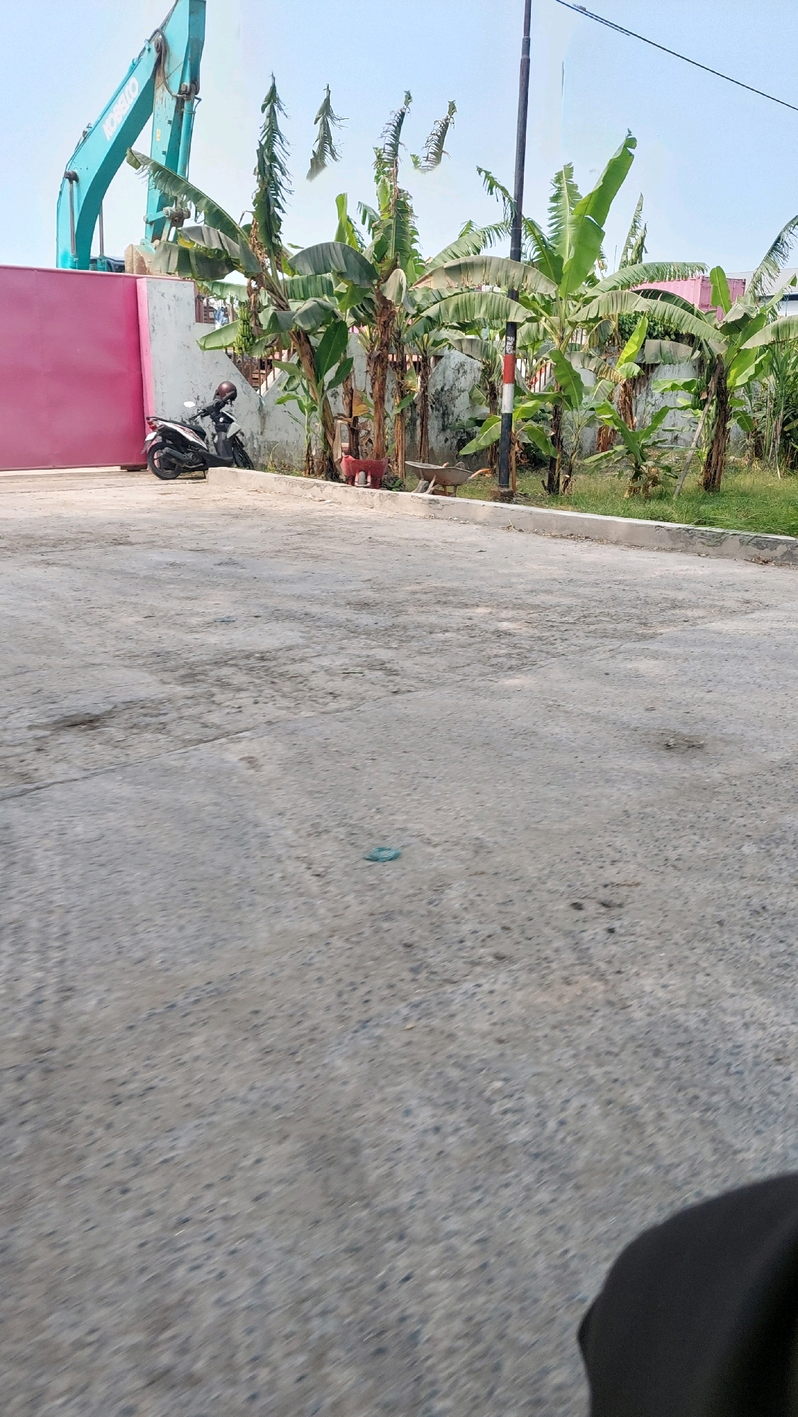 In Ketimang, Indonesia, a construction scene unfolds with a large teal excavator at the rear of a pink wall. In front of the wall, a black and white motorcycle is parked, with a helmet resting on its seat. Banana trees line the area, with some fronds appearing dry and frayed. A red, stylized animal sculpture stands near the banana trees, adjacent to a rustic wheelbarrow. A vertical pole with red and white markings stands prominently, with a black cable extending upwards. The ground is a rough, grey concrete surface, with some cracks and stains visible. The sky above is a pale blue.