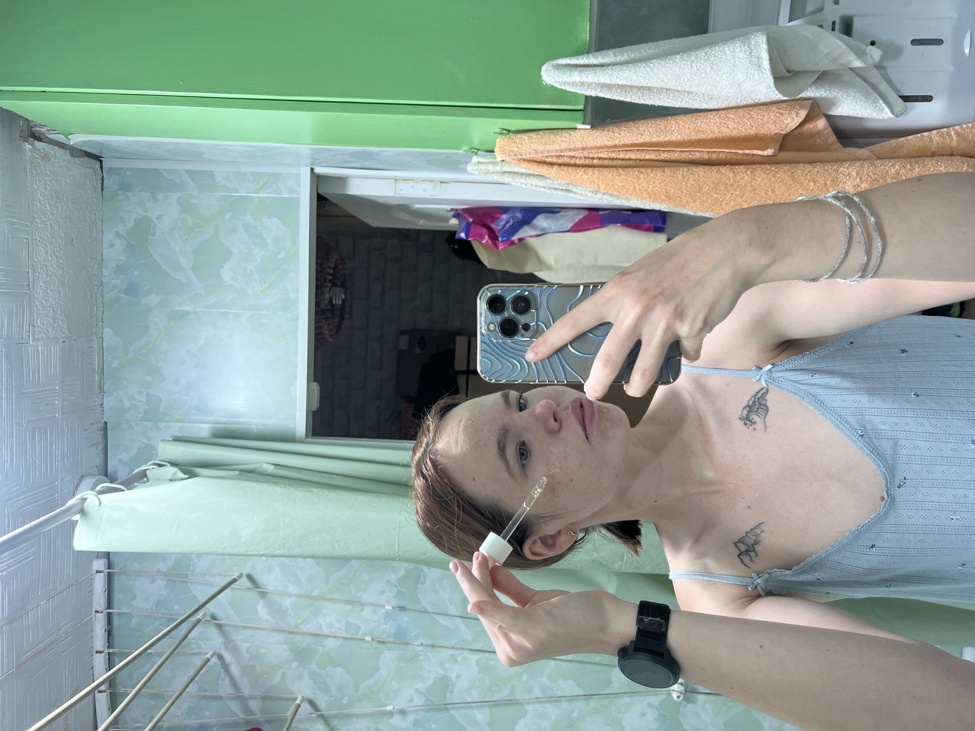 The image shows a young woman with light brown hair, wearing a light blue slip dress and a black watch on her left wrist. She is holding a dropper with a clear liquid to her cheek and taking a selfie with a smartphone. She has two tattoos on her chest: a mountain range on the left and a wave on the right. She is also wearing a bracelet on her right arm. The background appears to be a bathroom with light green tiled walls, a light green curtain, and several towels hanging on the right. The overall lighting is dim. The scene is consistent with a private bathroom in a residential building in Chaikovsky, Russia.
