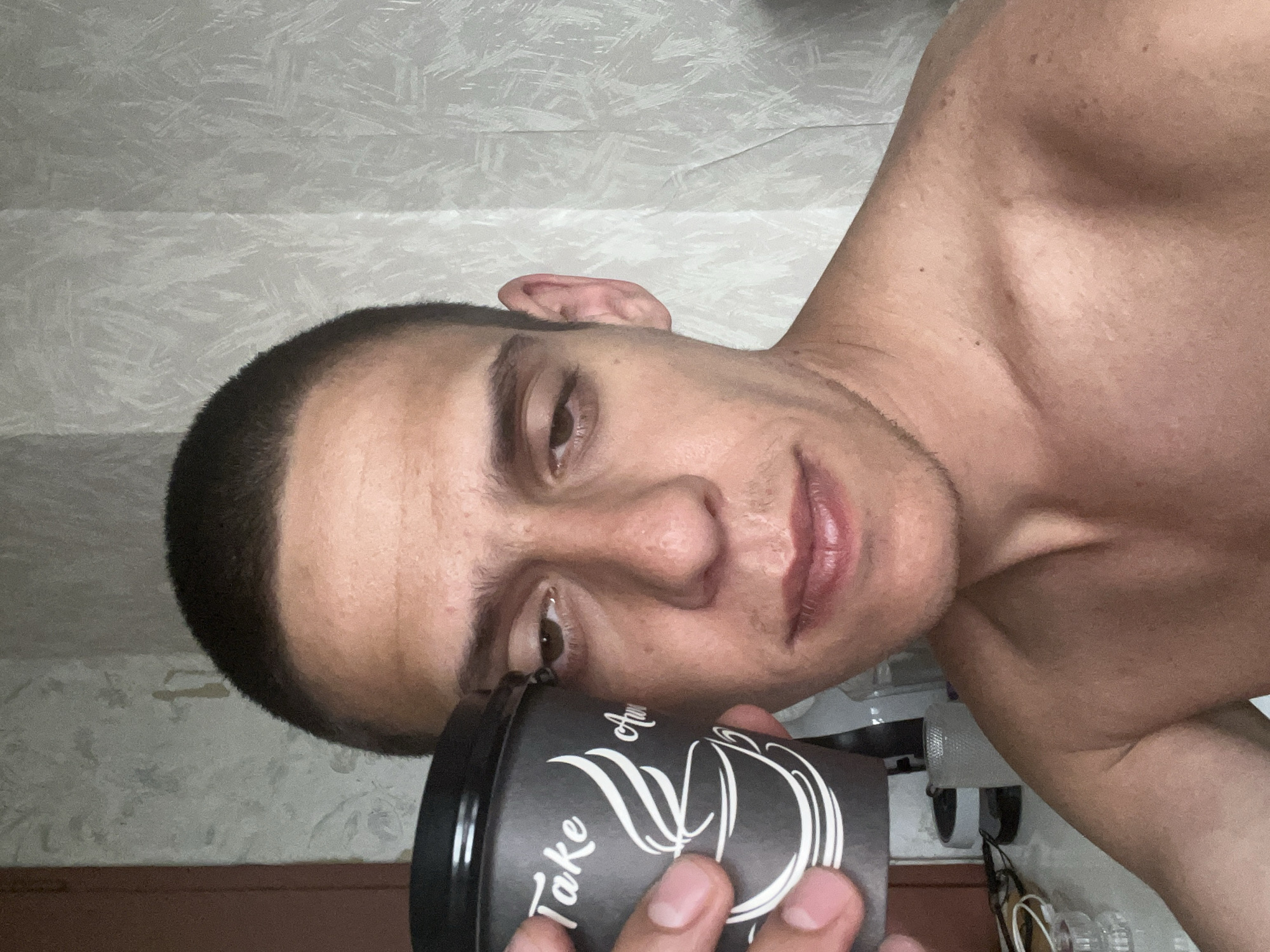 A shirtless man with short, dark hair holds a black cup with the word "Take" and a white swirl design. The background appears to be a room with textured wallpaper. The location in Sofia, Bulgaria is not discernible from the provided image.