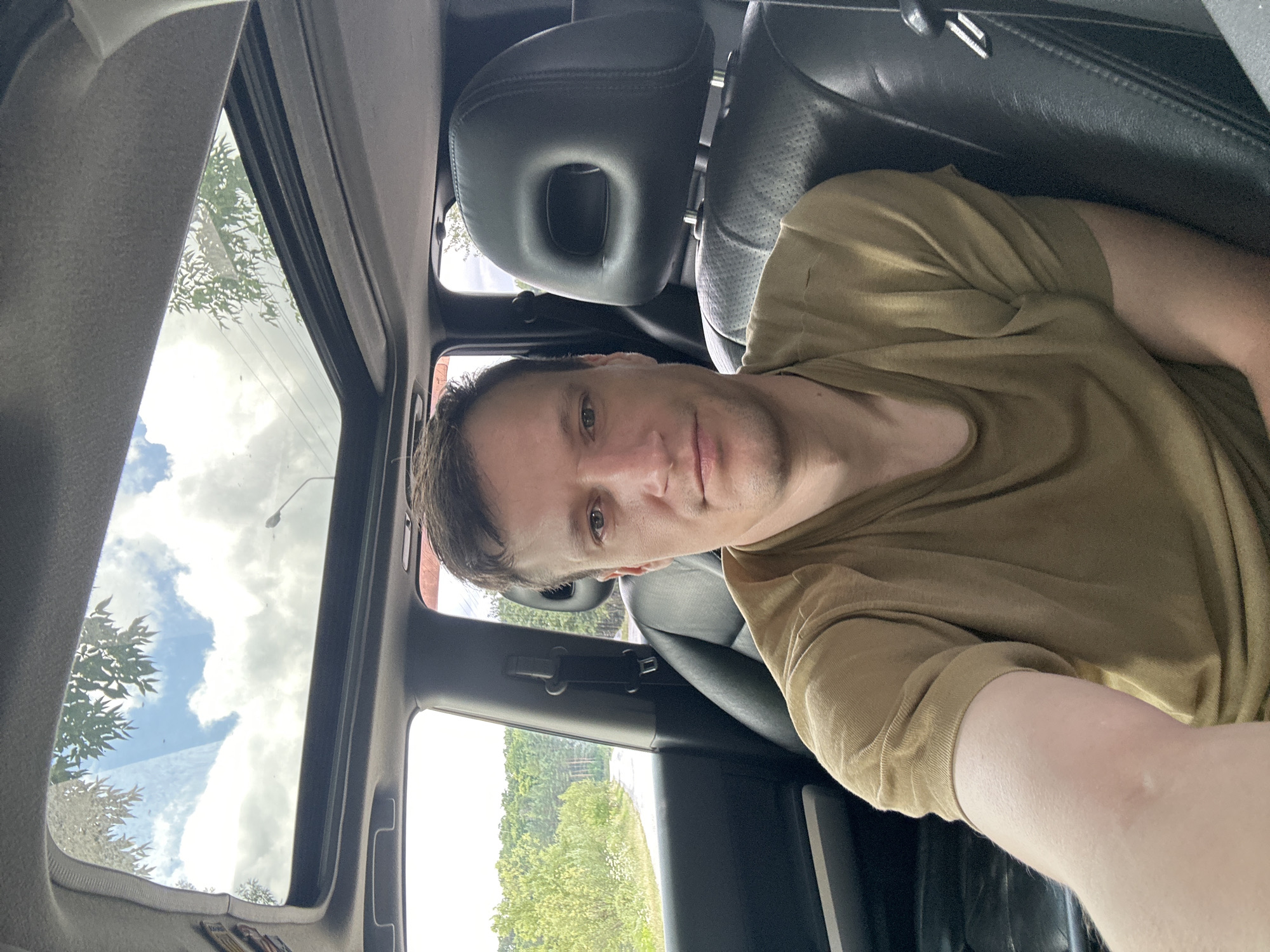 A selfie taken from the passenger seat of a car shows a Caucasian male with short brown hair and light skin, wearing a tan t-shirt. He is looking directly at the camera with a neutral expression. The car's interior is visible, with black leather seats and a sunroof open to a bright sky with clouds and trees. The exterior view through the windows shows a road winding through a green, wooded area, suggesting a rural or semi-rural setting. The scene appears to be set in Makiv, Ukraine, based on the provided information.