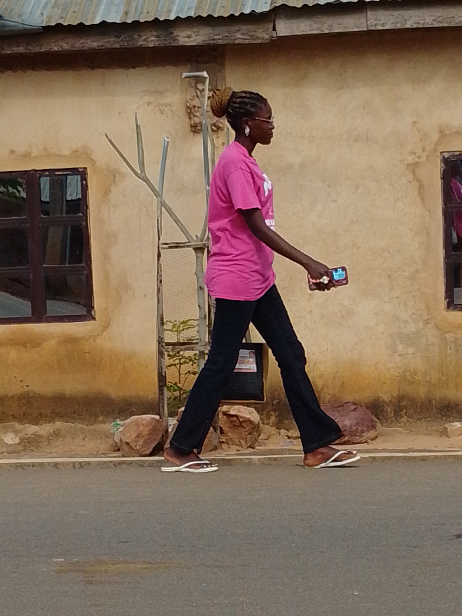 A medium shot captures a young Black woman walking from left to right, her body angled in profile. She is wearing a vibrant pink t-shirt with white writing on it and dark blue flared pants, paired with brown and white flip-flops. Her braided hair is pulled up, and she wears glasses and large white earrings. In her right hand, she holds a cell phone. The background consists of a light yellow, textured building with a corrugated metal roof. Two dark-framed windows are visible on the building, one on the left and another on the right, both reflecting some ambient light. In front of the building, a thin, weathered tree with sparse branches stands near a metal grate enclosure. Scattered rocks lie along the base of the wall and beside the paved street where the woman is walking. The scene appears to be an outdoor urban environment, possibly in Gombe, Nigeria, given the architectural style and vegetation.