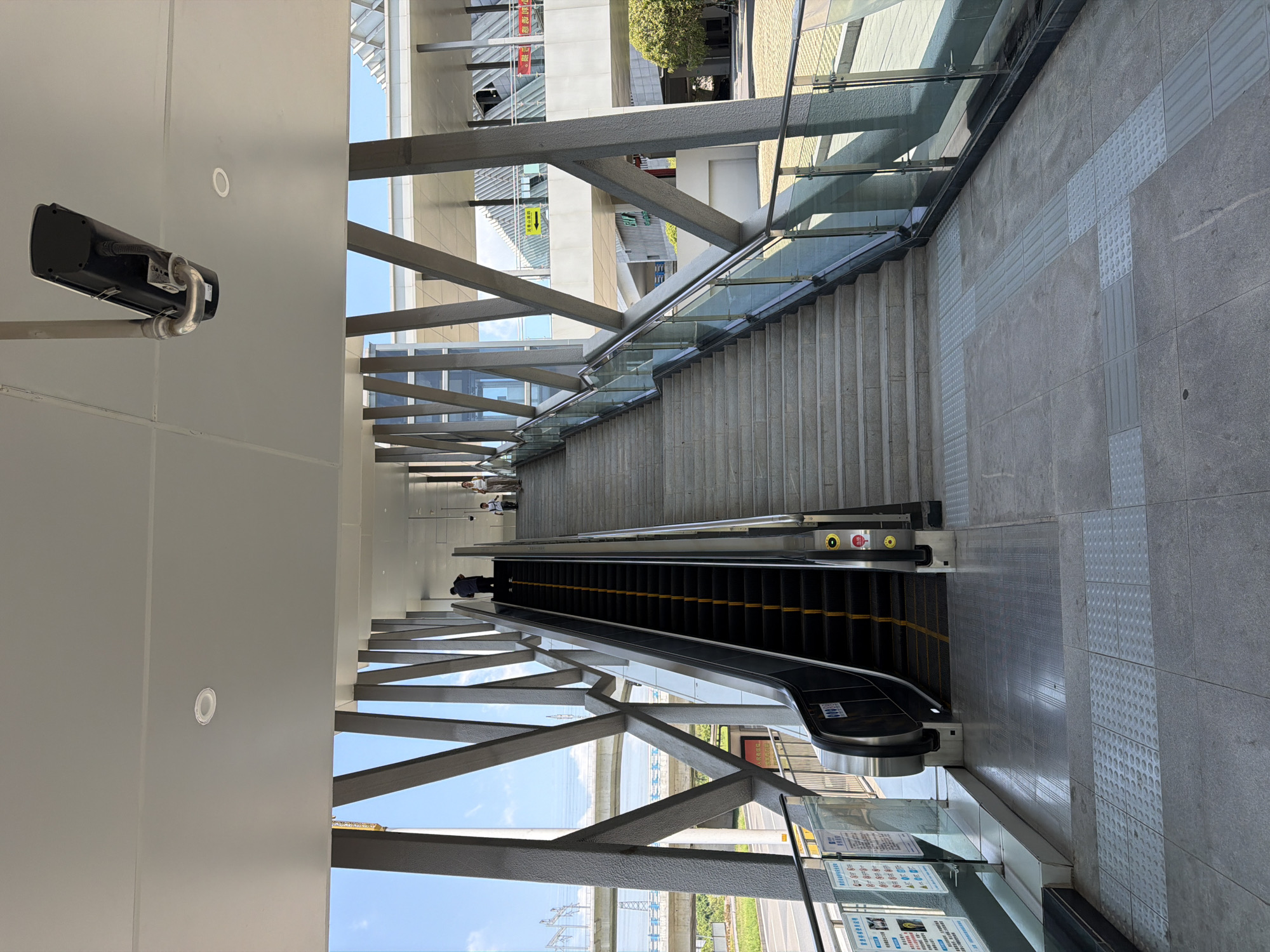 The image depicts an outdoor pedestrian overpass in the city of Jiangjin, China. The overpass features both an escalator and a set of stairs leading upwards. The structure is supported by a framework of angled metal beams. A security camera is mounted on the underside of the overpass ceiling. At the bottom of the escalator, there is a set of tactile paving tiles for the visually impaired. Two individuals are visible on the stairs, one ascending and the other further up. The sky is blue with some clouds. In the background, there are elevated railway tracks and buildings. The ground surface is paved with large gray tiles.