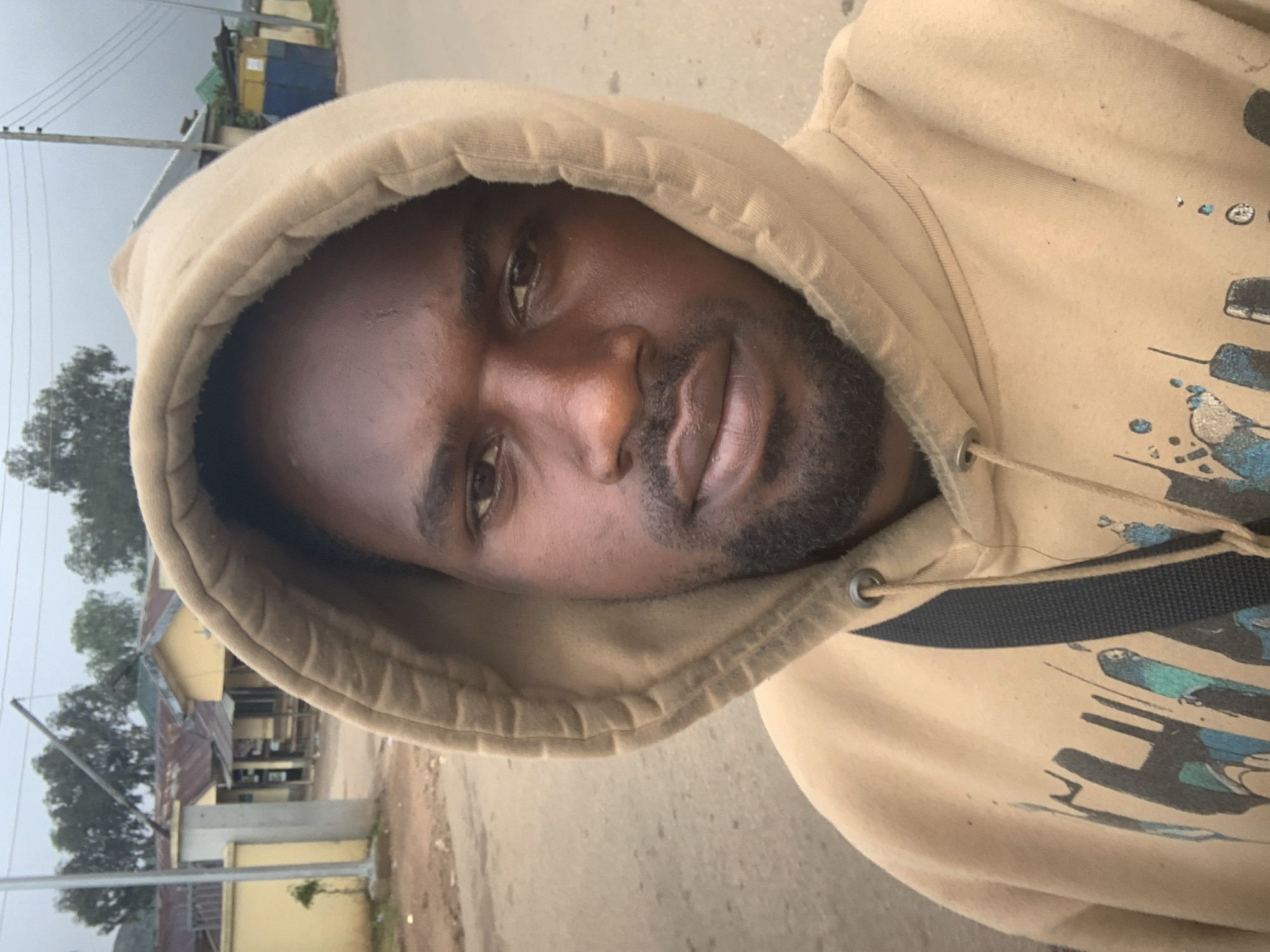 A selfie is taken from the chest up, showing a Black male with short dark hair and a beard, wearing a tan hooded sweatshirt. The hoodie is pulled up, framing his face. He has dark brown eyes, a prominent nose, and full lips. A black strap is visible diagonally across his chest, partially obscured by the hoodie. The background is an outdoor scene. To the left, a light-colored building with a red-tiled roof and windows can be seen. Beyond that, several trees are visible. To the right, a structure with a blue and yellow facade is present, along with power lines against a cloudy sky. The ground appears to be a paved road or street. The overall setting suggests a location in a residential or semi-urban area within Gura Top, Nigeria.