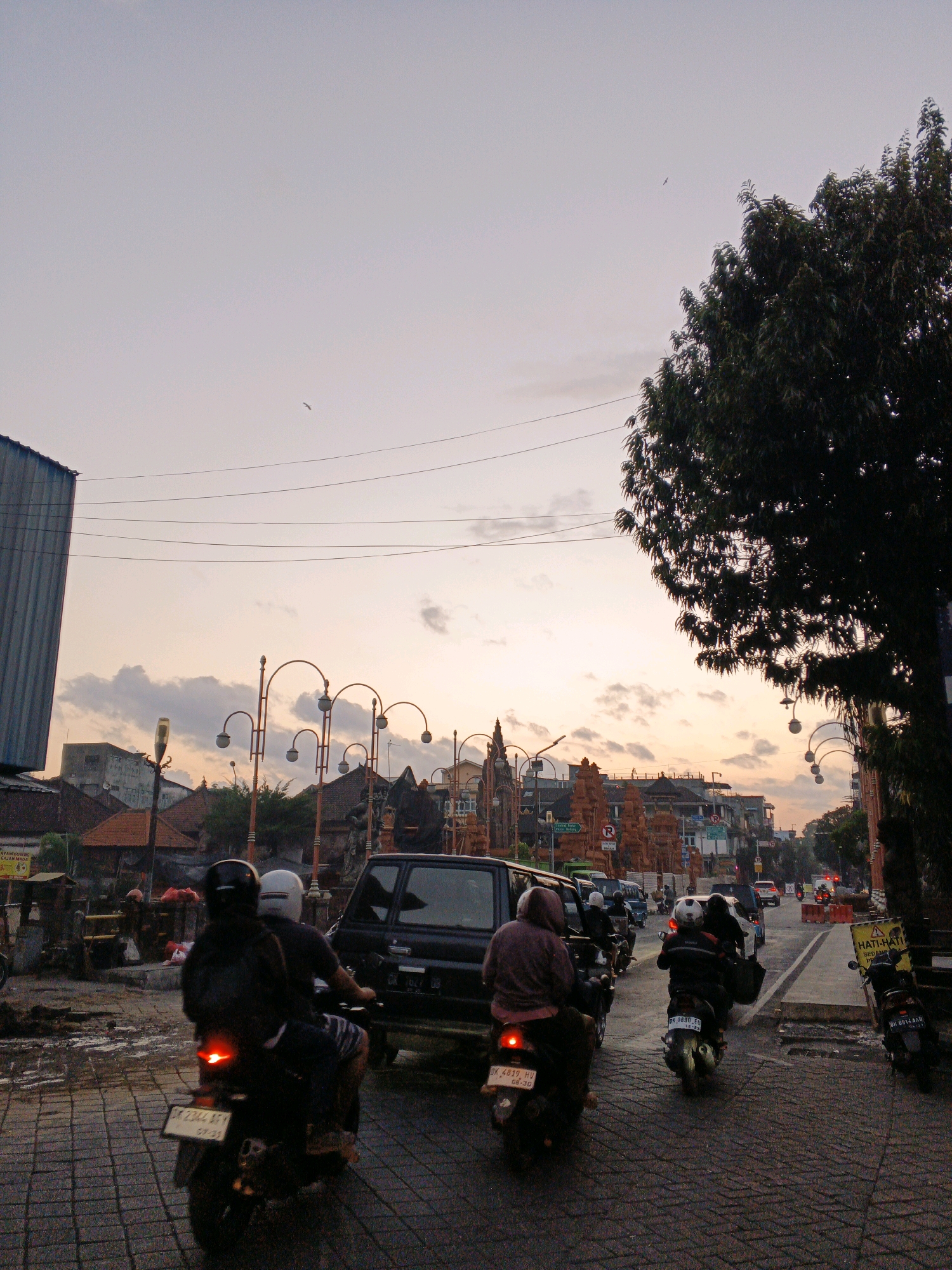 The image depicts a street scene in Denpasar, Indonesia, likely during the golden hour before sunset, given the warm hues in the sky. The sky is a pale orange and pink, with scattered clouds, and a single bird is visible soaring. Power lines crisscross the sky above a street paved with cobblestones.
In the foreground, several motorcycles are navigating the road. The closest motorcycle has two riders, both wearing helmets. The rider in front is wearing a black jacket and the rear rider wears a white helmet and a dark jacket. The motorcycle's brake light is illuminated. Behind them, another motorcycle with two riders, one wearing a light brown hooded jacket, is also visible, with its brake light on. Further down the street, more motorcycles and a black SUV are present in traffic.
On the right side of the image, a large, dark-leafed tree casts a shadow. On the left, a building with a corrugated metal exterior stands partially visible. In the background, traditional Balinese architecture is apparent with ornate gateways and buildings with tiled roofs. Streetlights with arched poles line the street. A sign with Indonesian text, "HATI-HATI SEDANG PECAH," which translates to "Be careful, being broken," is posted on the right side, accompanied by a warning symbol. The overall impression is of a busy urban environment at dusk.