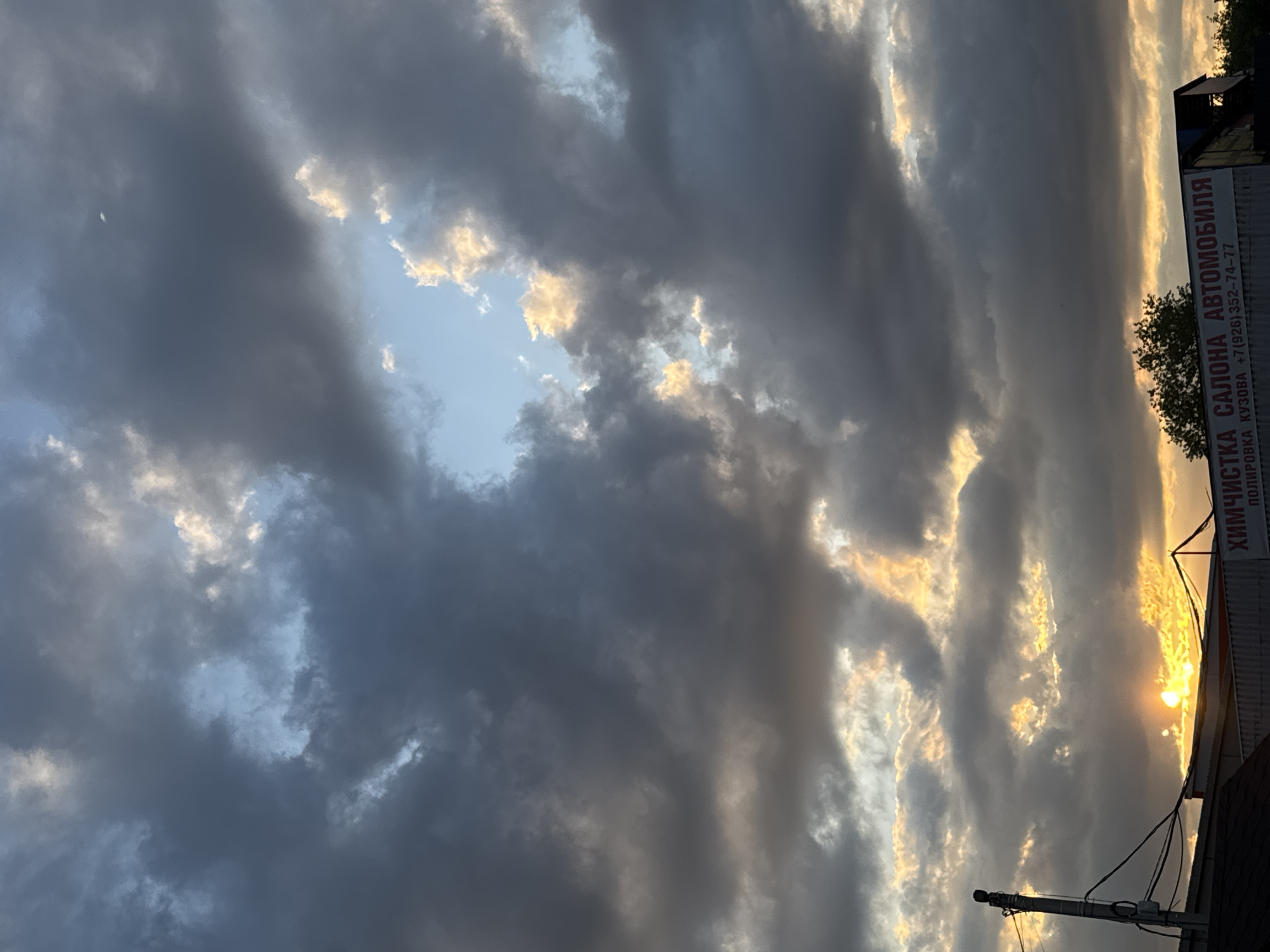 This is an eye-level, outdoor, medium shot of a dramatic cloudy sky at sunset, taken in Sovkhoza "Ramenskoe", Russia. The sun is setting behind a building with a visible sign that reads "ХИМЧИСТКА САЛОНА АВТОМОБИЛЯ ПОЛИРОВКА КУЗОВА" which translates to "Car Interior Cleaning, Body Polishing". A utility pole with wires is visible on the left. The clouds are dark and stormy, with patches of sunlight breaking through, creating an orange and yellow glow. The overall mood is ominous yet beautiful. There are no people or animals visible in the image.