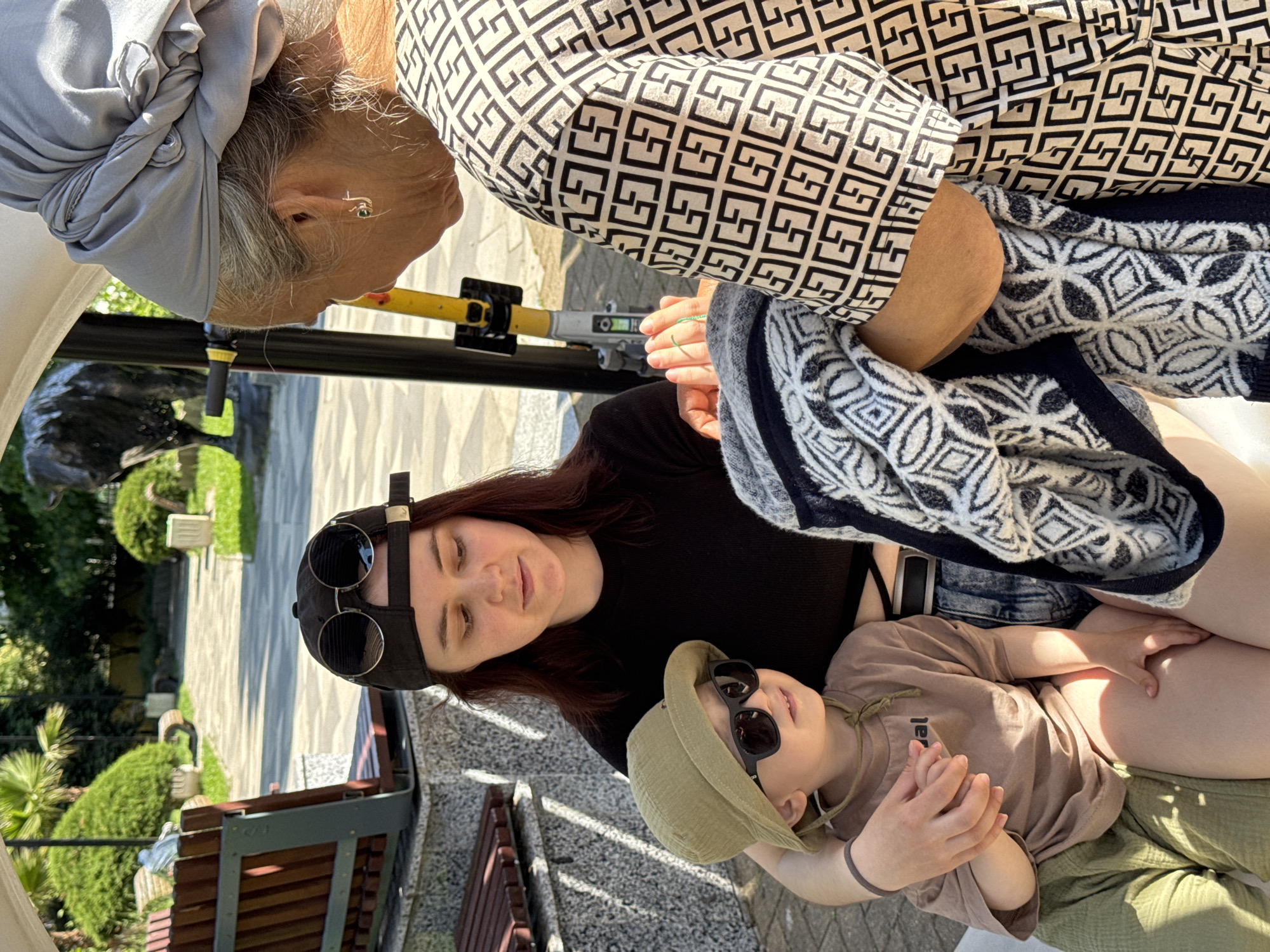 The image shows three people in an outdoor setting, possibly a patio or veranda with a view of greenery, suggesting a garden or park. The location is Sochi, Russia.
An elderly woman is positioned at the top, partially visible, wearing a headscarf and a patterned top. Below her is a younger woman with long hair, wearing a cap with sunglasses resting on top. A young child, wearing a hat and sunglasses, is seated on the younger woman's lap.
The background shows plants and a structure, possibly a bench, indicating an outdoor setting. The lighting suggests it is daytime.