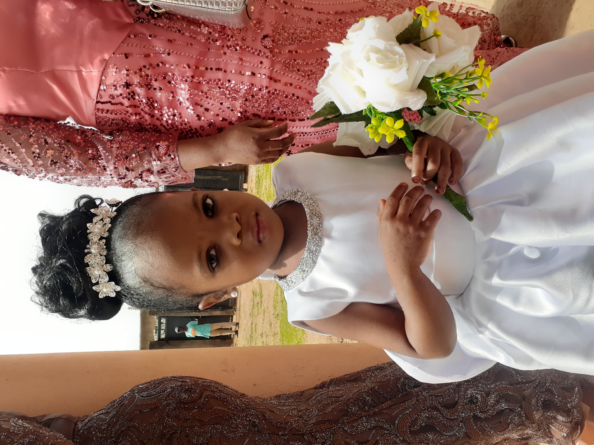 An upright, eye-level shot captures a young girl and a partially visible adult, likely at a formal event during daylight hours.
The primary subject is a young girl with dark skin, positioned centrally in the foreground. She gazes directly at the viewer with a serious, calm expression. Her black hair is styled in an updo, adorned with an elaborate silver and pearl floral hairpiece. She wears pearl stud earrings. The girl is dressed in a pristine white, short-sleeved dress with a satin-like texture. The high neckline features intricate silver, jewel-like embellishments. Her small hands, with red nail polish, delicately hold a bouquet of white roses and small yellow accent flowers.
Behind and to the right of the girl, the upper torso and arm of an adult are visible. This individual is wearing a long-sleeved, pink or rose gold dress, richly embellished with sequins. The bodice appears to be made of a plain, shiny fabric, contrasting with the sequined sleeves. One of the adult's hands is resting near the girl's shoulder.
The background suggests an outdoor setting. A warm-toned, earthy-brown structure or wall occupies the lower portion of the frame. Beyond this, a patch of green grass is visible, and in the distance, a person wearing a light blue shirt stands near what appears to be a doorway or gate. The overall impression is that of a special occasion, possibly a wedding, with the girl potentially serving as a flower girl. No text is visible in the image.
