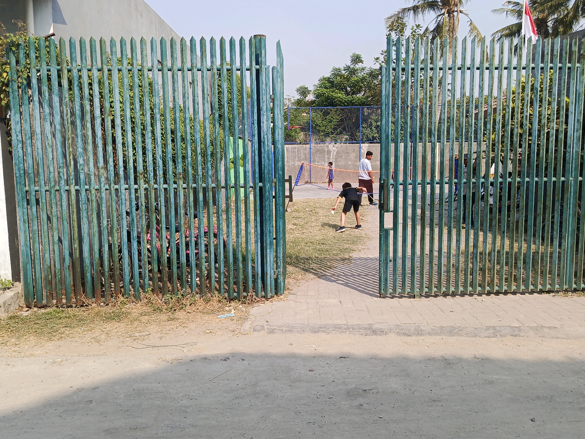 The image depicts an open gate in Jati Mulya, Indonesia, revealing a badminton court within a fenced-in area. Two young boys are engaged in playing badminton. One boy, wearing a black t-shirt and shorts, is in the foreground, actively hitting the shuttlecock. Another boy, in a blue t-shirt and shorts, stands further back on the court. An adult male, wearing a white t-shirt and pants, is also present near the net. The court is fenced with a blue net, and behind it, a concrete wall and trees are visible. The area outside the gate is a dirt and gravel path. The overall lighting suggests a bright, sunny day.