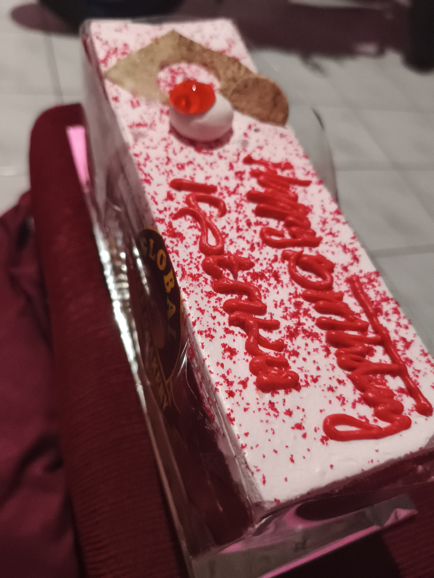 A close-up, slightly angled shot shows a rectangular cake with white frosting and red sprinkles, placed on a maroon cloth surface. The cake is decorated with red icing that forms cursive lettering on top, appearing to say "Happy Birthday." A red jelly-like topping and a small piece of tan confectionery are also present on the cake. A portion of a circular label with the word "GLOBAL" is visible on the side of the cake. The background is out of focus and appears to be indoors with a soft light source. There are no people visible in the image. The location in Depok, Indonesia, is not discernible from the visual information provided.
