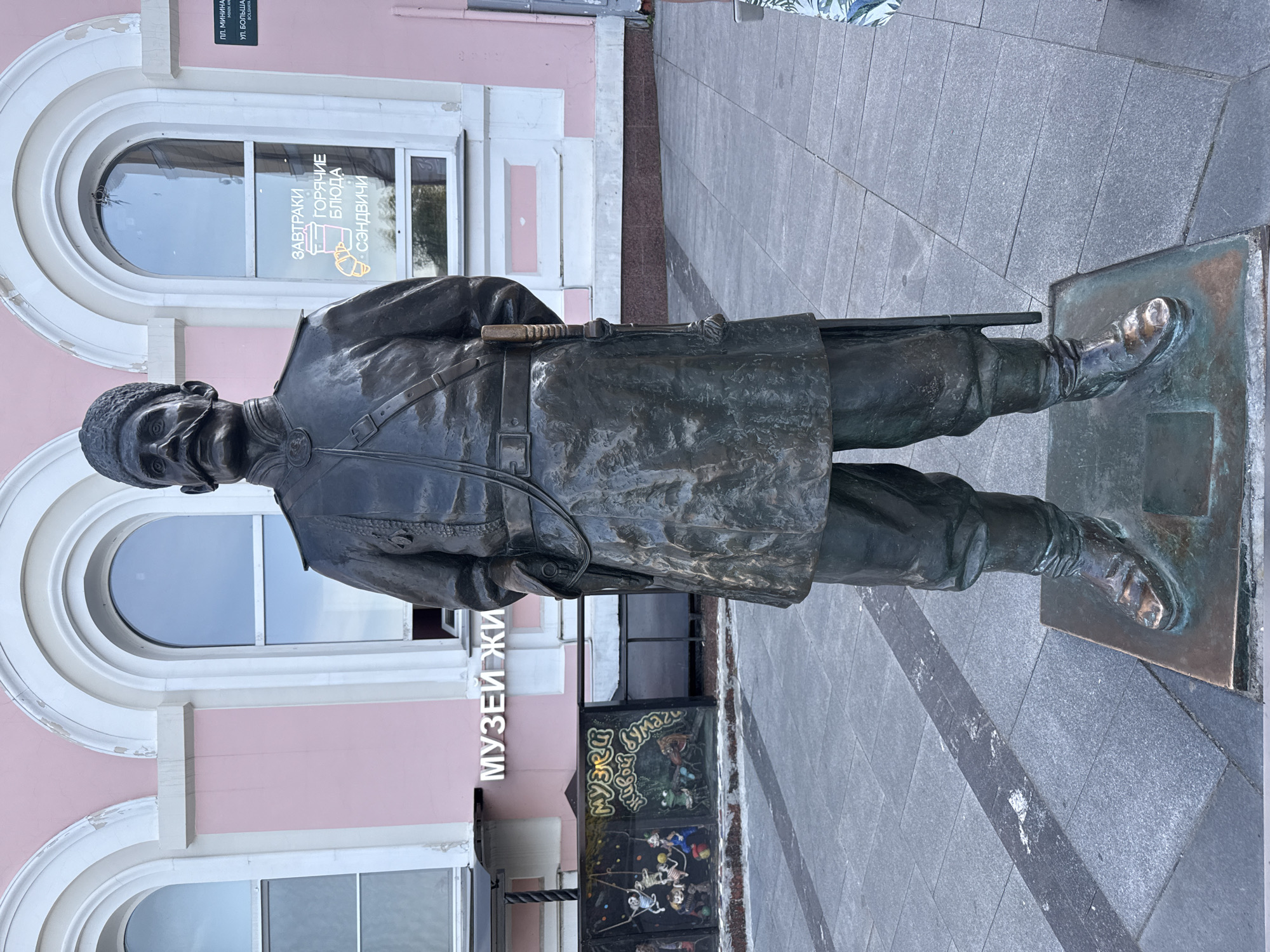 A full-body bronze statue of a man in military attire stands on a stone sidewalk in front of a pink building. The man is facing forward and has a mustache and a hat. He is wearing a long coat, pants, and boots. A sword is sheathed at his hip. A sign with Russian text is visible on the building behind him, reading "МУЗЕЙ ЖИ". Other signs with Cyrillic text are visible on the building. To the left of the statue, a black panel with colorful illustrations is displayed. The sidewalk is made of gray stone pavers. The scene is outdoors, and the lighting suggests it is daytime. The location is Nizhny Novgorod, Russia.