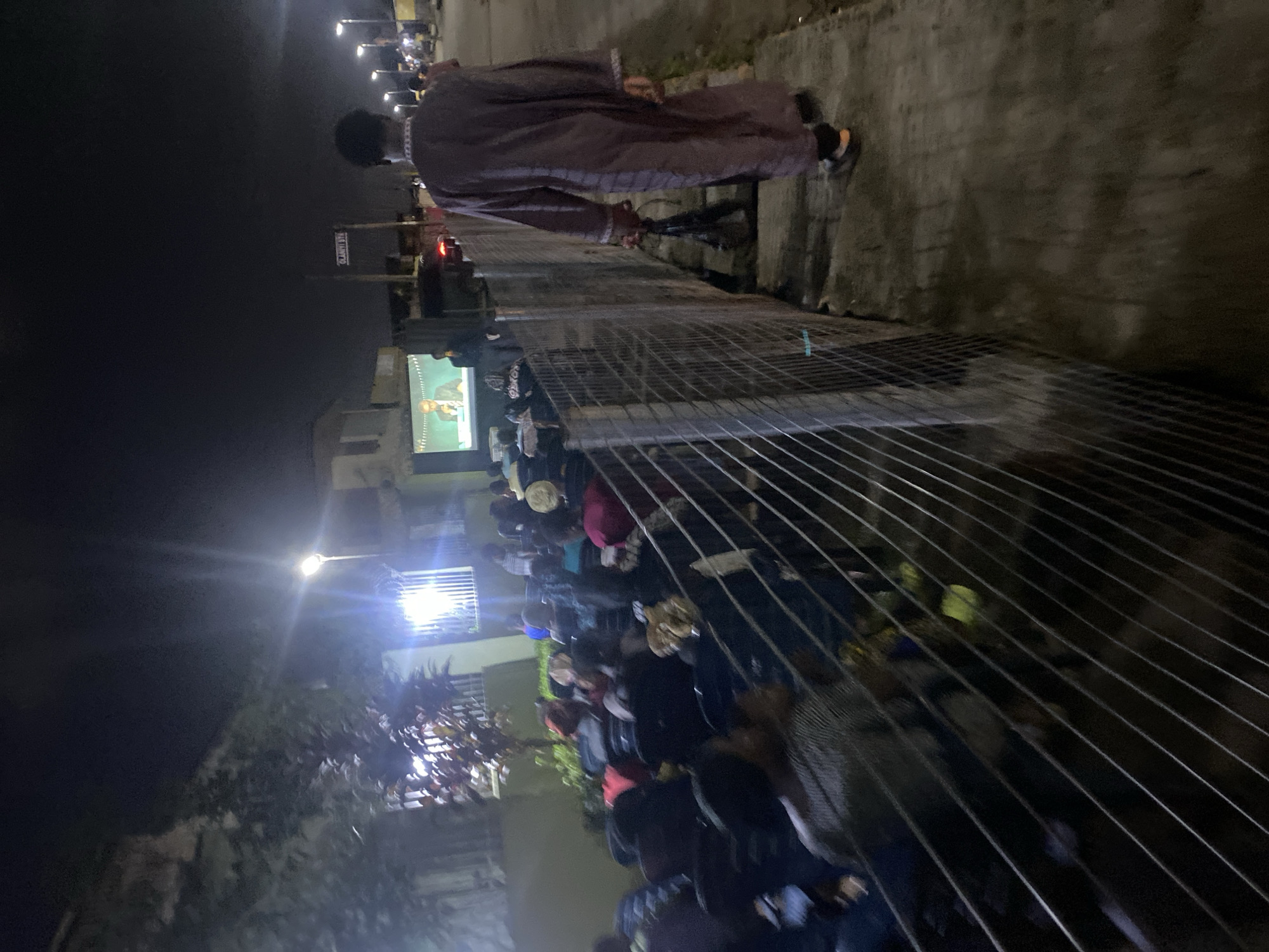 At night in Lagos, Nigeria, a large crowd has gathered outdoors, likely to watch a projected video, possibly for entertainment or a public event. A man in a long, patterned robe stands near a fence, seemingly observing the crowd or the video. The environment is urban, indicated by buildings, street lights, and the fence. The lighting is dim, but a screen is visible among the gathering.