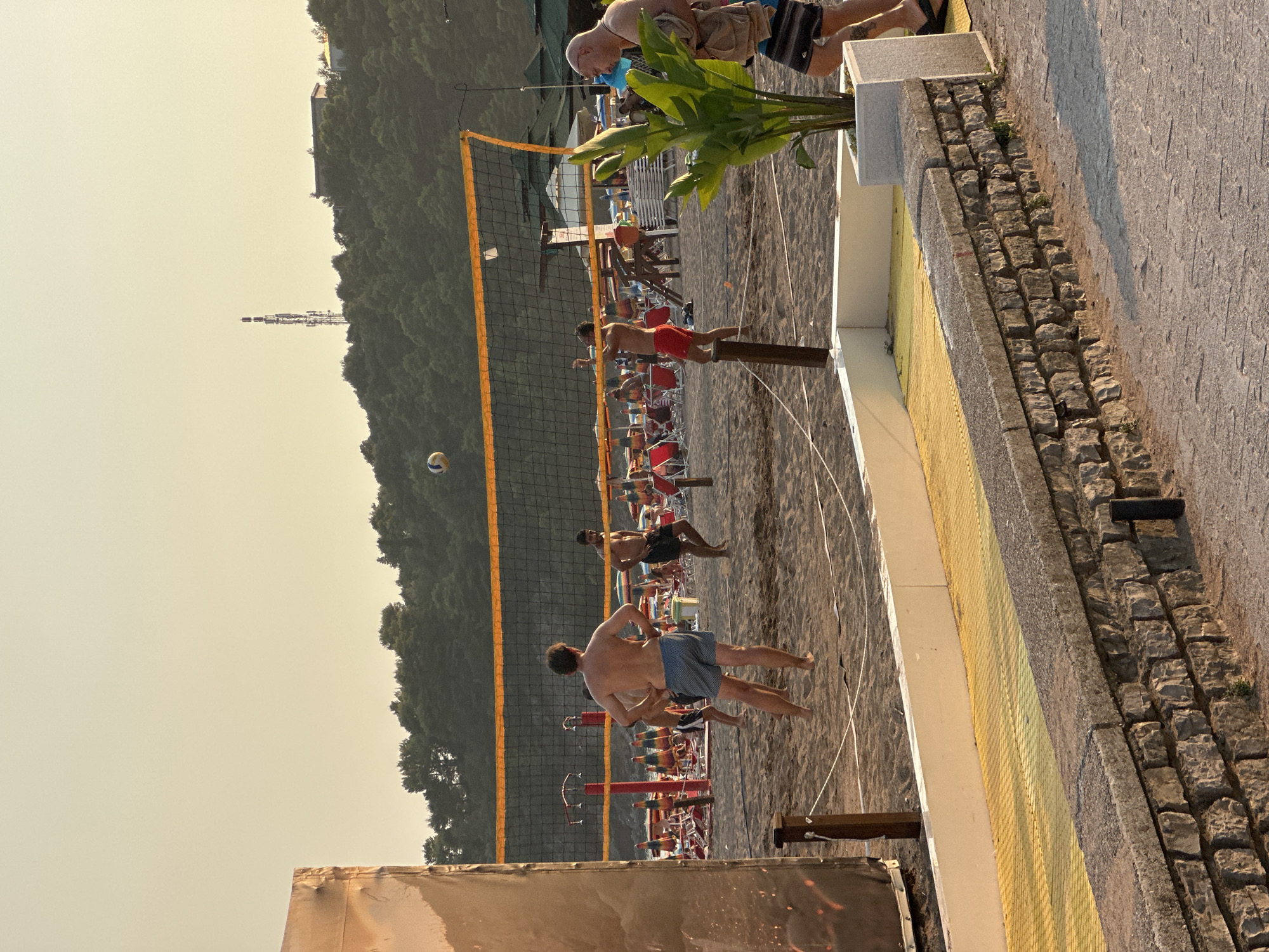 In Boreti, Montenegro, on a sandy beach, a group of men are playing a game of beach volleyball. A volleyball is visible in mid-air above the net. The game is taking place near a line of beach chairs and umbrellas. The sun is setting, casting a warm glow over the scene. The image is taken from a slightly elevated perspective, showing a stone wall and a yellow walkway in the foreground.