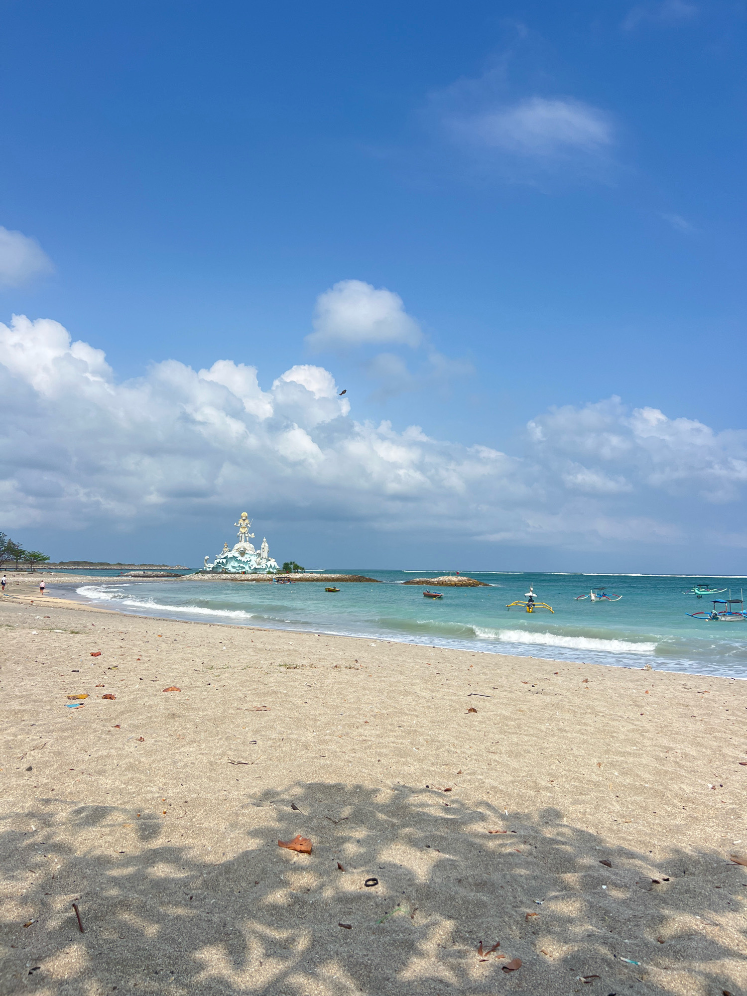 This image shows a beach in Kuta, Indonesia. The sand is light brown and there are some small pieces of debris scattered on it, including dried leaves and plastic. The ocean is turquoise and there are gentle waves breaking on the shore. In the distance, there is a large white statue of a seated figure with elaborate golden headdress, standing on a rock formation in the water. Several traditional Balinese fishing boats, with outriggers and colorful decorations, are visible in the sea. The sky is blue with scattered white clouds. Two individuals are walking on the beach in the background, and a bird is flying in the sky. The foreground of the image is dominated by the sandy beach with shadows from nearby trees cast across it.