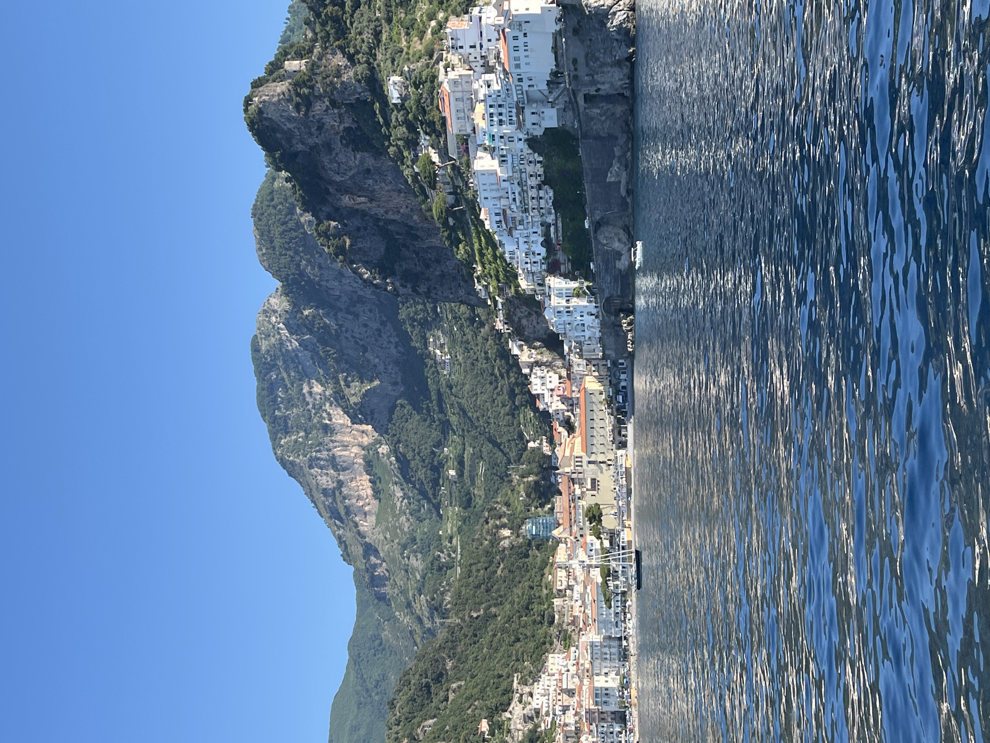 A scenic view of the Amalfi Coast in Pogerola, Italy, showcases the picturesque town nestled against lush, mountainous terrain. White buildings with red-tiled roofs cascade down the steep slopes towards the azure sea. The water in the foreground is a deep blue, rippling with sunlight, reflecting the vibrant sky. A sailboat with dark sails is anchored in the bay, and a small white boat is visible near the shore. The backdrop is dominated by rugged mountains covered in dense green vegetation, with terraced slopes suggesting agricultural activity. The sky above is a clear, bright blue, indicating a sunny day.