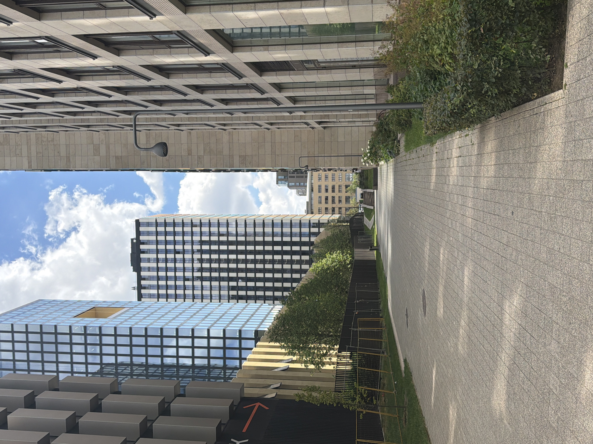 The image shows a modern street scene in Moscow, Russia, with contemporary architecture and landscaping. On the left, a building with a dark facade and distinctive metal panels is visible, next to a tall glass skyscraper with a geometric pattern of windows and a prominent orange square accent. To the right of this, another tall building features a black and white checkered facade. In the center background, a lower building with a grid of windows extends towards the horizon, and further back, another building with a uniform facade can be seen. The right side of the image is dominated by a building with a light-colored, stone-like facade, featuring rows of tall windows and a modern street lamp. A paved pathway, made of light-colored rectangular stones, runs through the scene, bordered by green grass and neatly trimmed bushes. A fence separates the pathway from the grass and trees on the left side. The sky above is blue with scattered white clouds. There are no people or animals visible in the image. The overall impression is one of urban development and contemporary design.