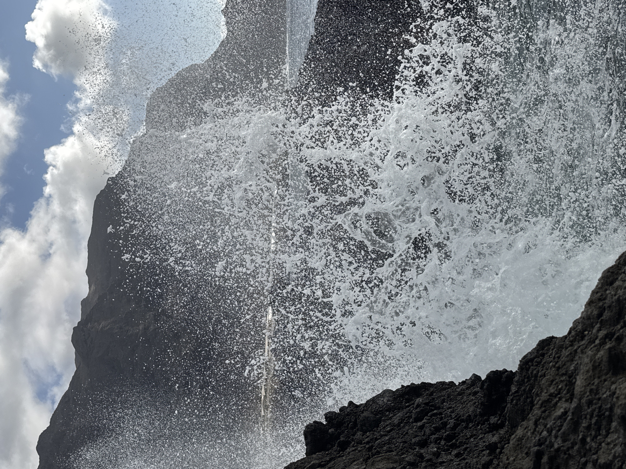 An outdoor, close-up, eye-level, vertical shot shows a wave crashing against dark, rocky cliffs under a partly cloudy, bright blue sky. The splash of water fills most of the frame with a spray of white foam and droplets. In the upper portion of the image, dark, mountainous terrain is visible, along with a sliver of the ocean and a distant horizon line. The scene is dynamic, capturing the raw power of the ocean.