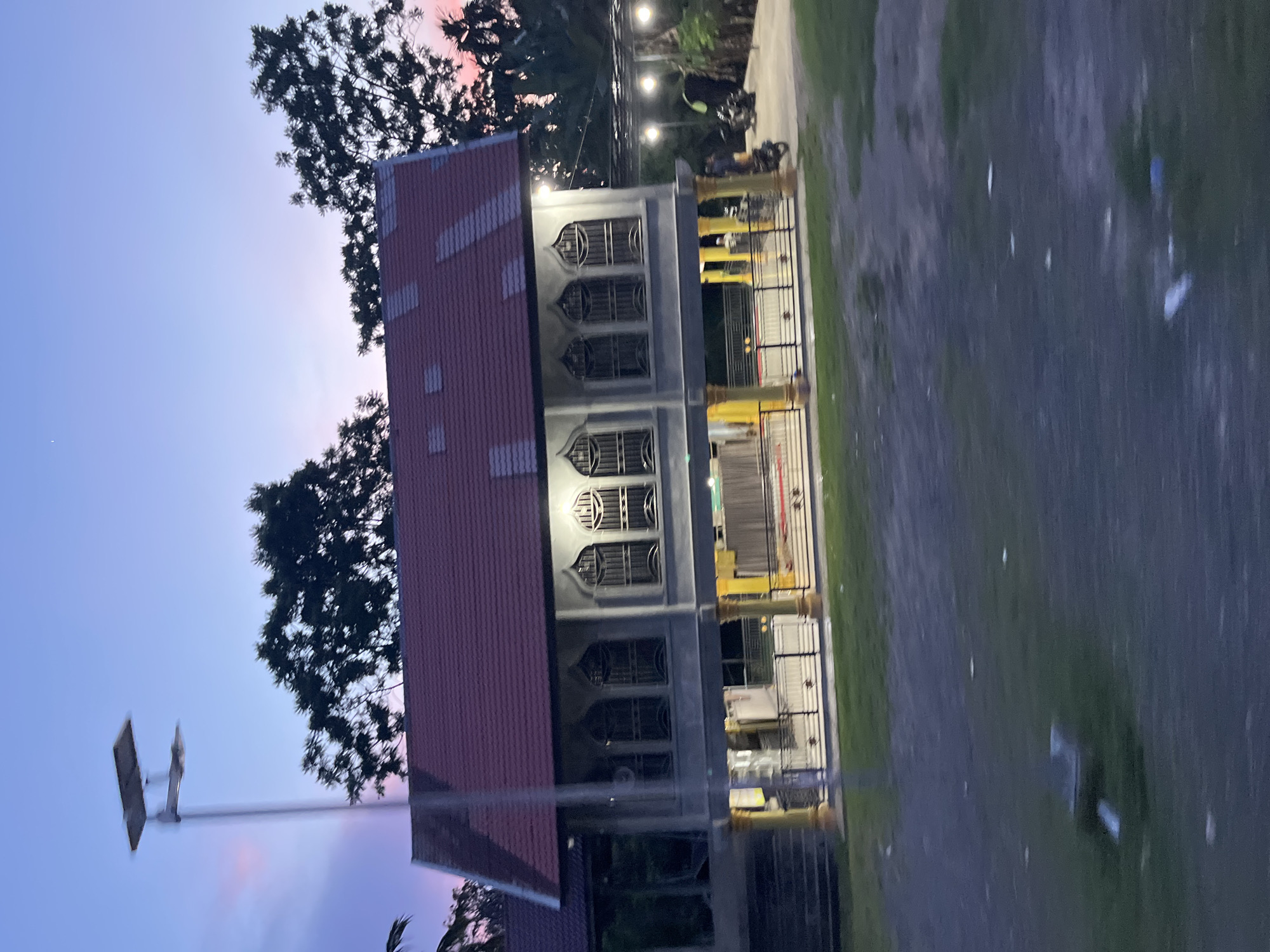 The image depicts a stilted building with a red tiled roof, reminiscent of a mosque or community hall, situated in Matangpanyang, Indonesia. The building is elevated on yellow and gray pillars and features multiple arched windows with intricate metalwork. The sky above displays a gradient of purple and pink hues, indicating twilight. A solar-powered street lamp stands to the left of the building. In the foreground, a grassy area is visible with some scattered debris. A black fence encloses the area around the building. To the right, a paved area shows two motorcycles and several outdoor lights. The overall scene is tranquil, capturing a moment at dusk in a rural or semi-rural Indonesian setting.