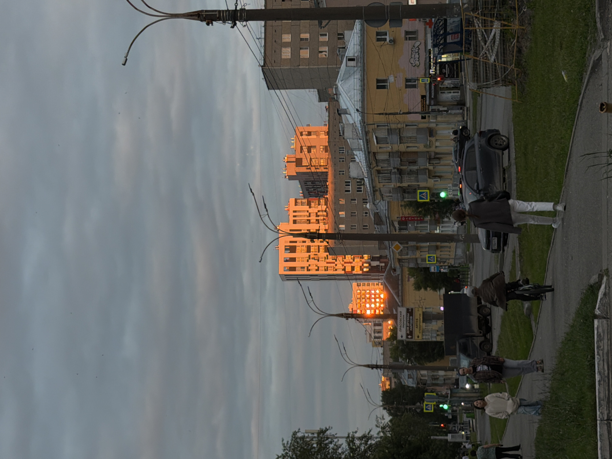 This image depicts a street scene in Izhevsk, Russia, during what appears to be sunset. The low angle of the sun casts a warm, golden light on the buildings, creating a striking contrast with the overcast sky. The scene is dominated by residential buildings, some of which show signs of age and wear.
Several individuals are present in the image, engaged in everyday activities. In the foreground, two young women stand on a sidewalk, one wearing a light-colored sweater and jeans, the other in a darker jacket and white pants. Further down the sidewalk, a person on a bicycle is visible. In the background, a dark-colored car is parked. Traffic lights are visible, with one showing a green signal.
Notable details include the distinct architectural style of the buildings, characteristic of Soviet-era construction. The street is lined with trees, and a grassy area runs alongside the sidewalk. The lighting suggests it is late afternoon or early evening.
Visible text includes street signs. One sign, pointing to the right, has Cyrillic text that includes "ПРАВОСЛАВНЫЙ" and possibly "ПРОСПЕКТ," indicating a street name and a religious building. Another sign has "HAРÓДН" which likely refers to "народный" (national/people's). There are also yellow pedestrian crossing signs with the Russian word "ПЕРЕХОД" (crossing). Graffiti is visible on some of the buildings.