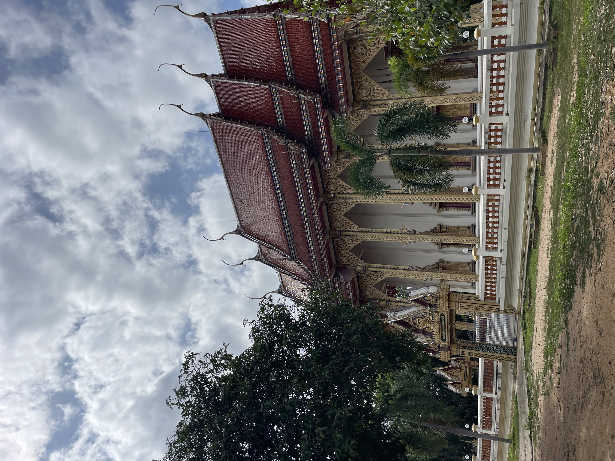 The image shows a traditional Thai temple in Pattaya City, Thailand. The temple features ornate architecture with multiple tiered roofs, decorative eaves, and golden embellishments. A large green tree is visible on the left side of the frame, with several palm trees in front of the temple structure. A fence with decorative railings runs along the front of the temple. The sky above is partly cloudy with a mix of white clouds and blue sky. The foreground consists of a dirt and grass area. No people are present in the image.
