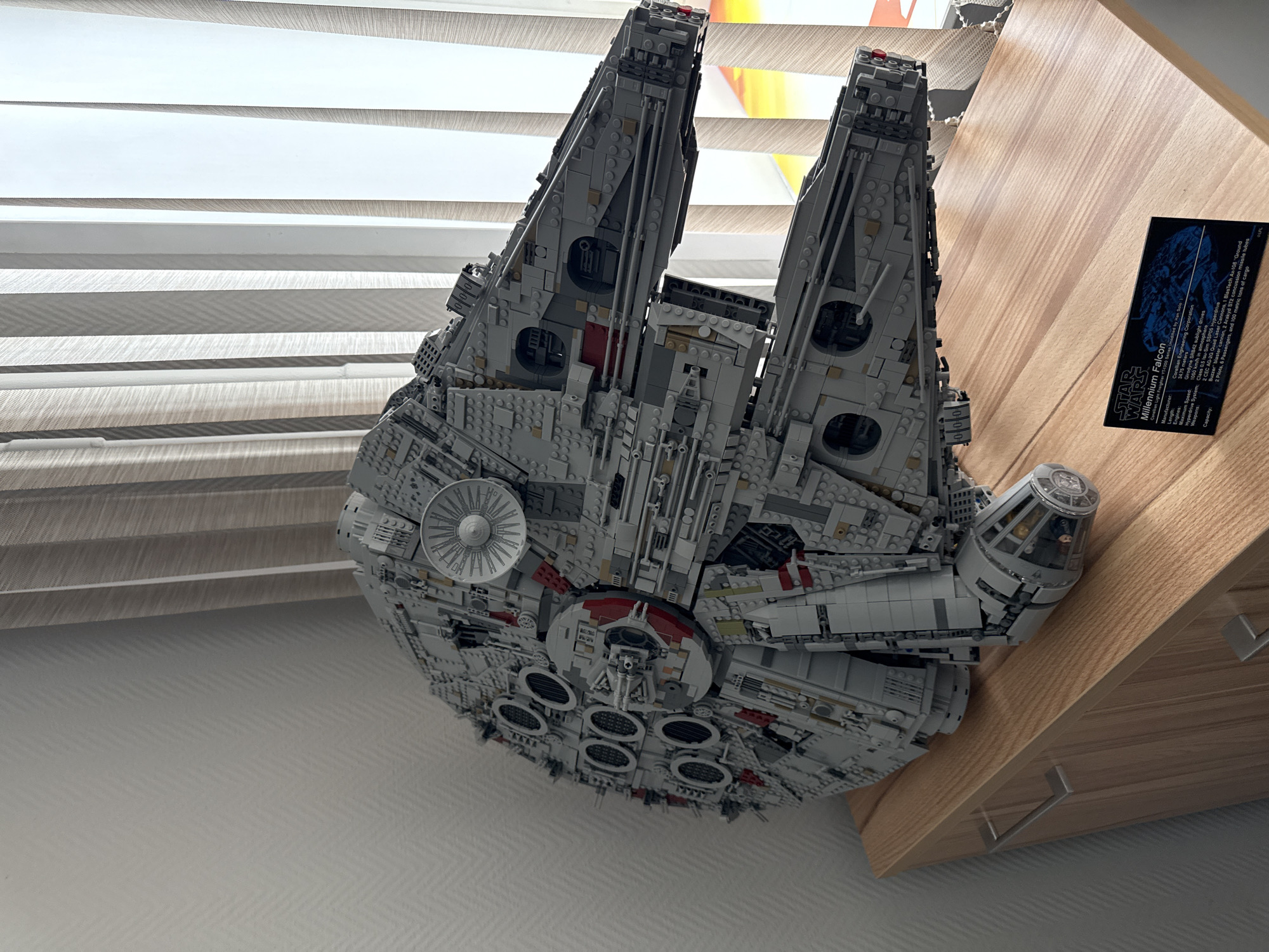 The image shows a detailed model of the Millennium Falcon from Star Wars. It's made of LEGO bricks. The model is displayed on top of a light-wood-colored cabinet or shelf. A placard with information about the Millennium Falcon is placed near the front of the shelf. The background includes a window with horizontally striped blinds. The image provides no information to indicate the location is in Krasnoyarsk, Russia.