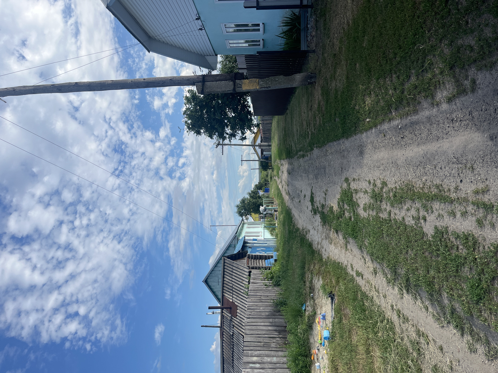 This is a vertical, eye-level, daytime photograph taken in Сухринское, Russia. The image captures a rural residential street scene. A dirt road runs diagonally from the bottom right to the top center of the frame, flanked by grassy areas and houses.
On the left side of the road, in the foreground, is a weathered, wooden house with a corrugated roof. A metal pipe protrudes from the roof. Further down the street, another house painted light blue with white trim is visible, featuring several windows. A dark fence separates this property from the road. A large wooden utility pole with wires stretches across the upper left portion of the image.
On the right side of the road, the grassy embankment is interspersed with patches of bare dirt. Further down the street, more houses and trees can be seen. In the far distance, the road continues into a cluster of trees. There are some plastic bottles and children's toys scattered along the edge of the road on the right.
The sky is bright blue with scattered white, puffy clouds, suggesting a clear and sunny day. The lighting indicates it is likely midday. The overall mood is tranquil and everyday, reflecting a quiet village life. No people are visible in the image. There is no visible text.