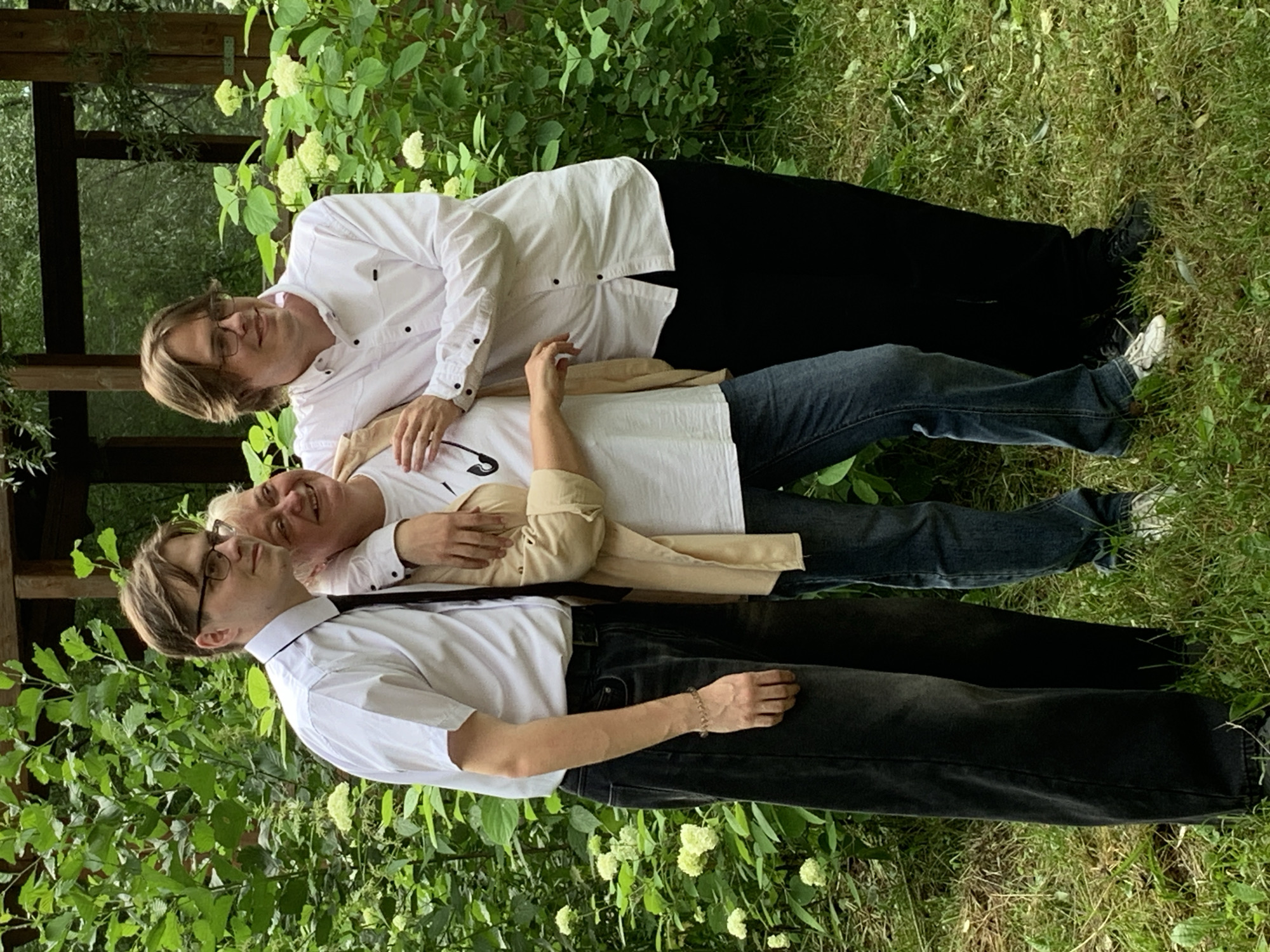 Three individuals are posing for a photo in an outdoor, natural setting, likely a garden or park, amidst lush green foliage and flowering bushes. The overall impression is a casual, familial gathering.
From left to right:
The first individual is a young man wearing glasses, a white collared shirt with a dark tie, and black jeans. He has a light complexion and light brown hair. He is standing and looking towards the right side of the frame. A thin, gold-colored bracelet is visible on his left wrist.
The second individual is a woman, positioned slightly in front and between the two men. She has blonde hair and is smiling broadly, looking towards the viewer. She is wearing a white t-shirt and a light-colored open jacket or cardigan. Her left arm is around the shoulder of the man on her left, and her right arm is resting on the shoulder of the man on her right.
The third individual is a young man, also wearing glasses, a white collared shirt, and black pants. He has light brown hair and is looking towards the left side of the frame, with a slight smile. His right arm is around the woman's shoulders.
The environment consists of dense greenery with scattered white flowers, suggesting a spring or summer season. The ground is covered in grass and some fallen leaves. In the background, a dark wooden structure, possibly a gazebo or part of a building, is visible. The lighting suggests it is daytime, with soft, diffused light.
There is no specific indication in the image to place the location in the city of Chaikovsky, Russia.