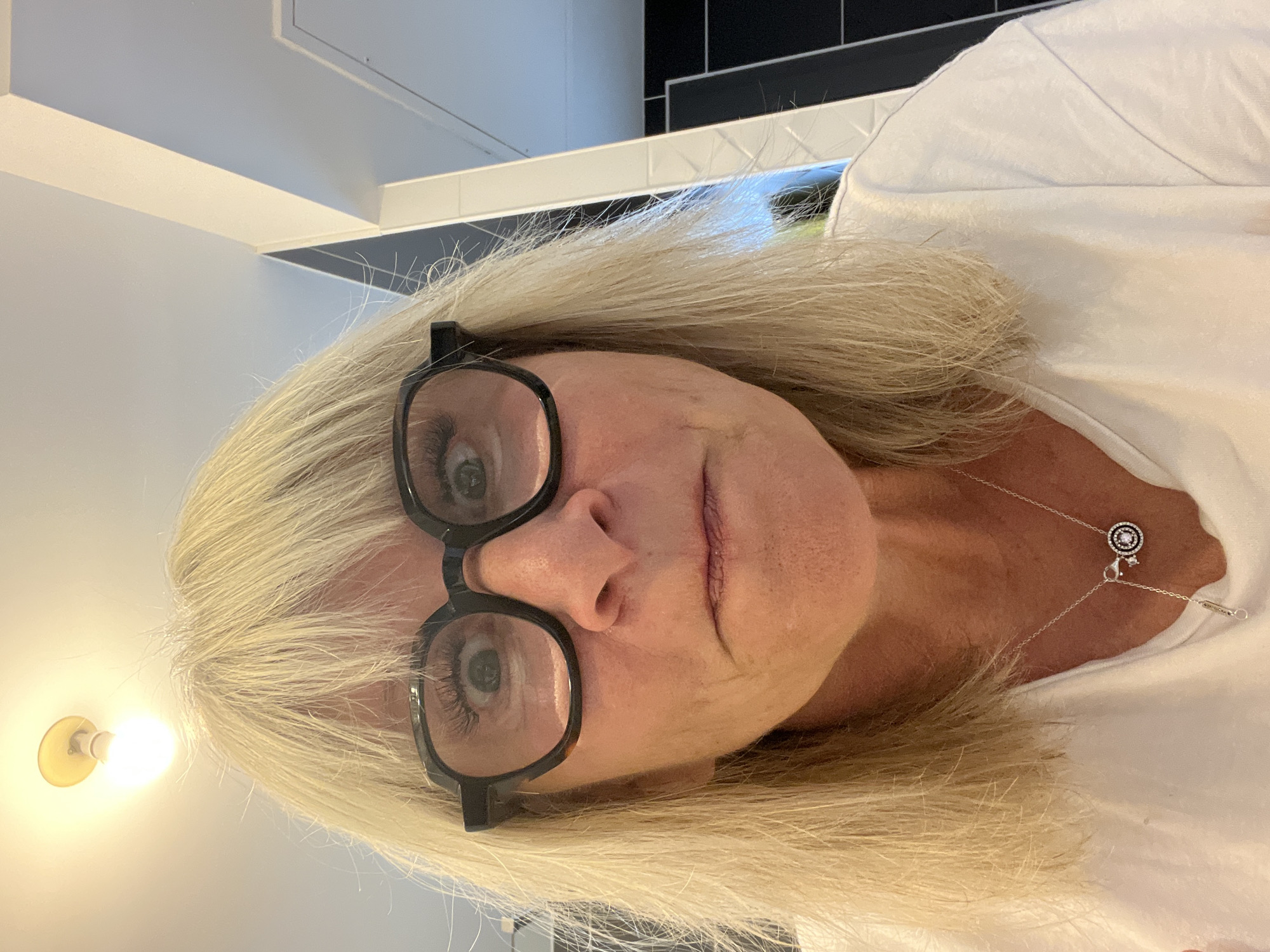 A close-up, medium-shot captures a middle-aged woman with blond hair and black-rimmed glasses. She wears a white shirt and a delicate silver necklace. The background includes a black and white tiled wall and a light fixture. The location is presumed to be Chambœuf, France.