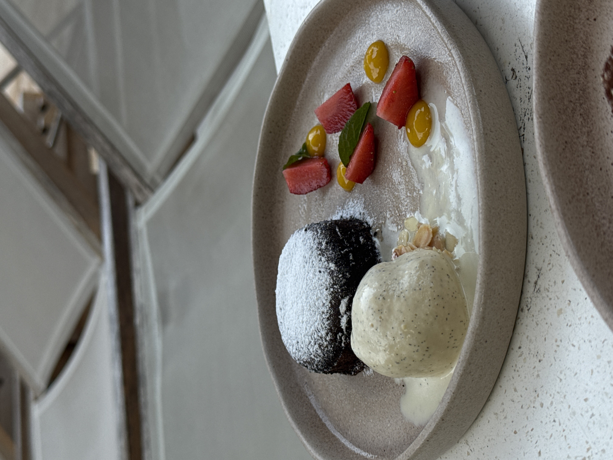 A close-up, eye-level shot displays a chocolate lava cake dusted with powdered sugar, served on a taupe plate. Accompanying the cake are a scoop of vanilla ice cream speckled with black seeds, sliced strawberries, a few mint leaves, and dollops of a yellow sauce, possibly mango or passionfruit. The dessert is arranged on a light-colored, speckled countertop. In the blurred background, two white lounge chairs are visible, suggesting an outdoor setting, possibly a patio or resort. The scene appears to be set in an unknown city, Unknown.