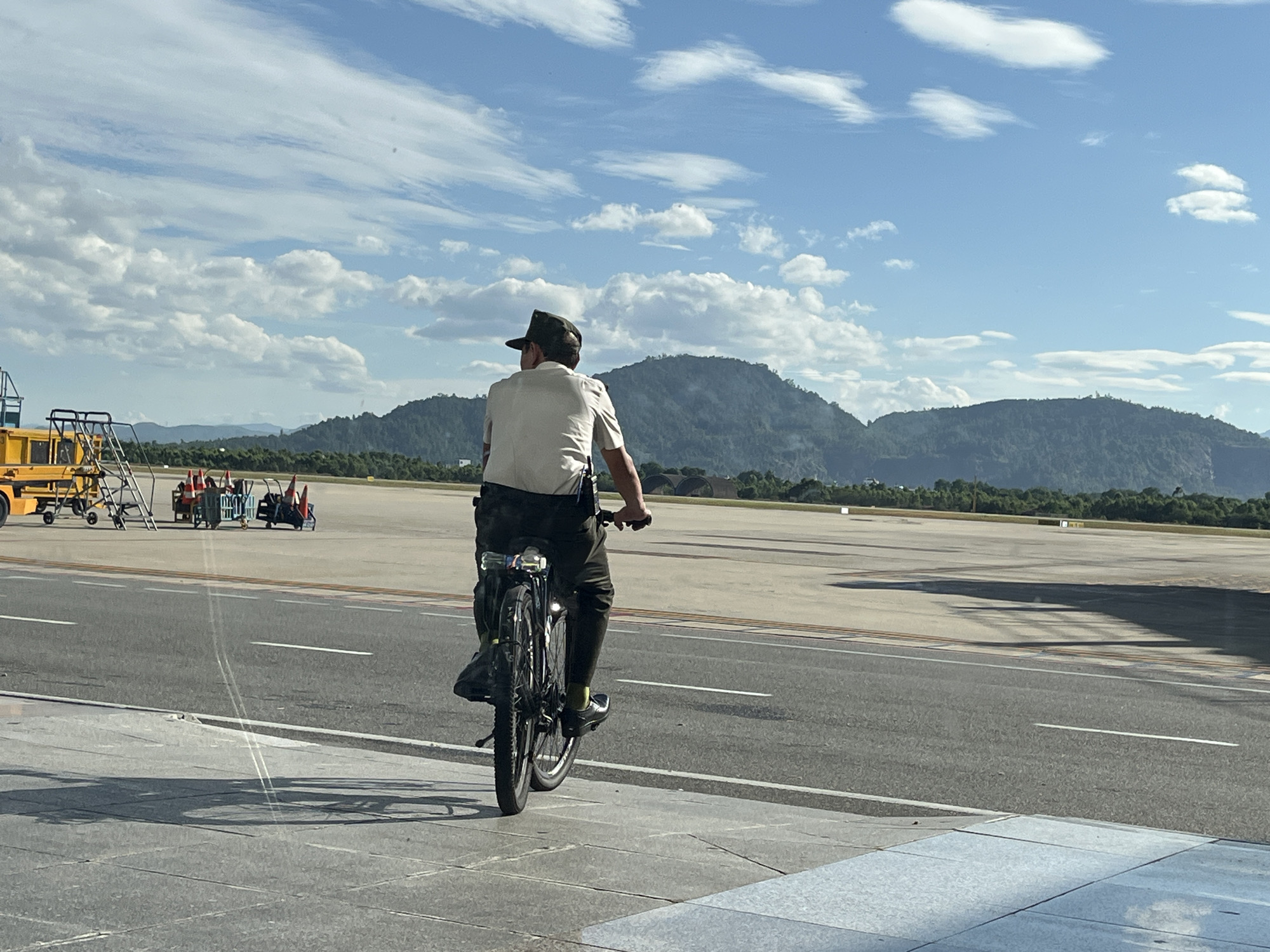 The image captures an outdoor scene at Da Nang International Airport in Da Nang, Vietnam, under a clear blue sky with scattered white cumulus clouds. In the foreground, a concrete sidewalk is visible. An adult male, viewed from behind, is riding a dark-colored bicycle on an asphalt road adjacent to the sidewalk. He wears a light-colored, short-sleeved collared shirt, dark trousers, a dark cap, and bright green/yellow socks with dark shoes. A radio or similar device is clipped to his right hip. The bicycle features a rear rack with a prominent rear light and possibly a water bottle or battery pack.
Beyond the road, a wide, light-colored concrete airport tarmac or runway stretches into the mid-ground. On the left side of the tarmac, various pieces of ground support equipment are present, including a yellow vehicle (possibly a passenger stair or conveyor belt loader) and multiple metal carts containing orange traffic cones. In the distant background, a range of lush, green, tree-covered mountains or hills rise under the sunny sky. Strong sunlight casts distinct shadows of the cyclist and bicycle onto the sidewalk and road surface.